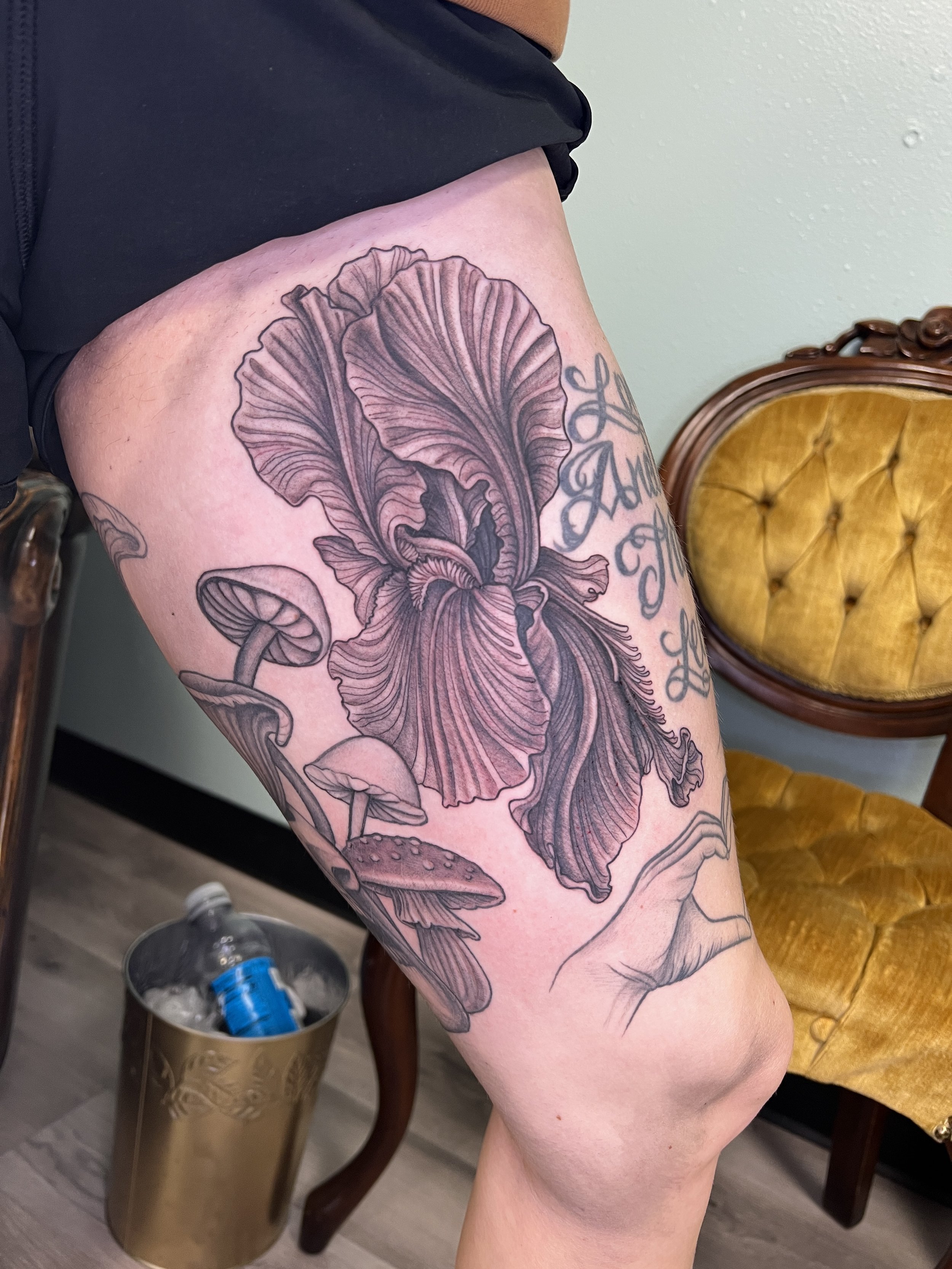 A black and gray tattoo of a large, detailed mushroom and iris on a person's upper thigh.