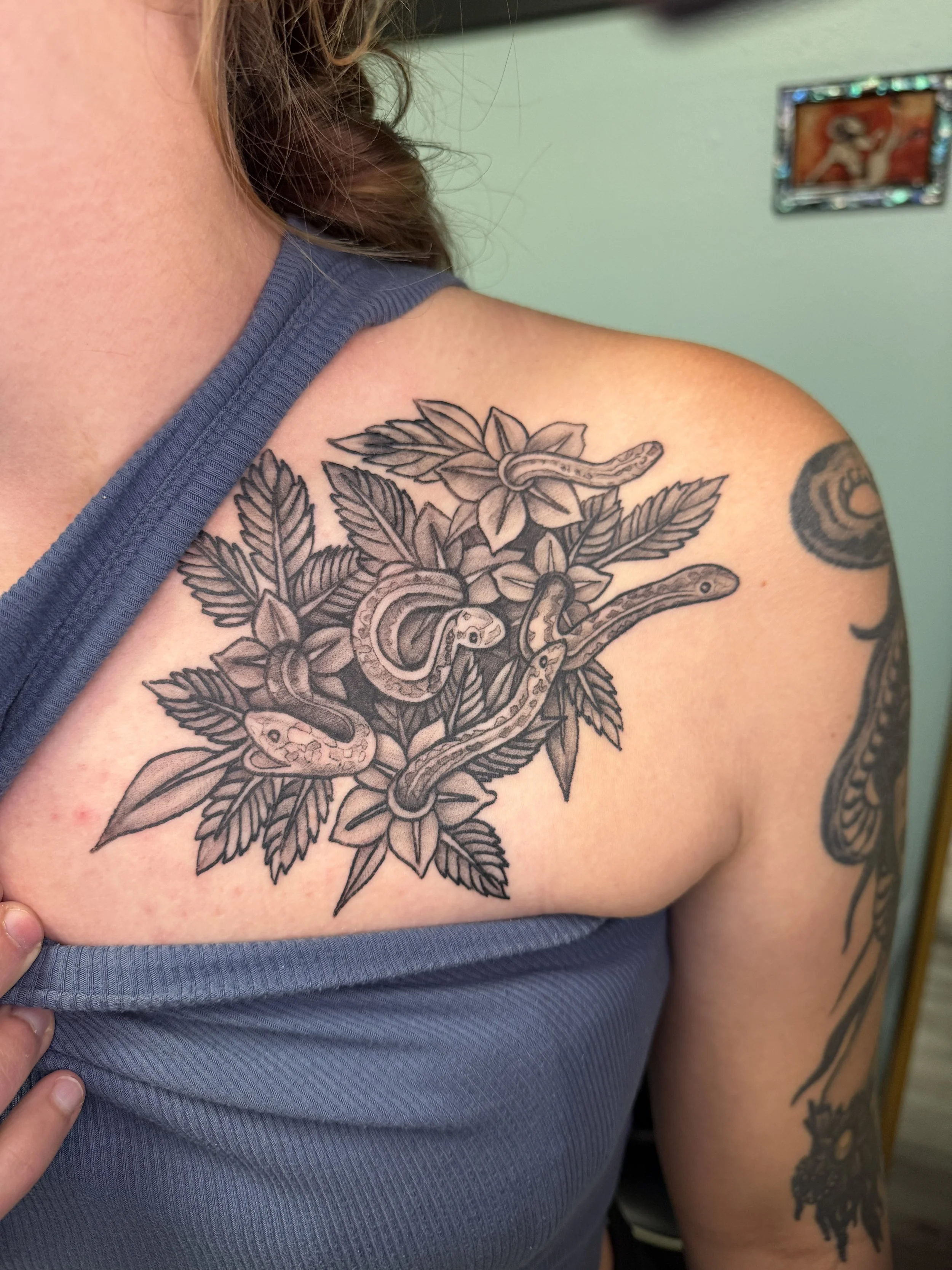 Close-up of a person's shoulder with a black ink tattoo of a snake coiled around a bouquet of flowers and leaves. The person is wearing a blue sleeveless top. There is a small colorful picture frame on the wall in the background.