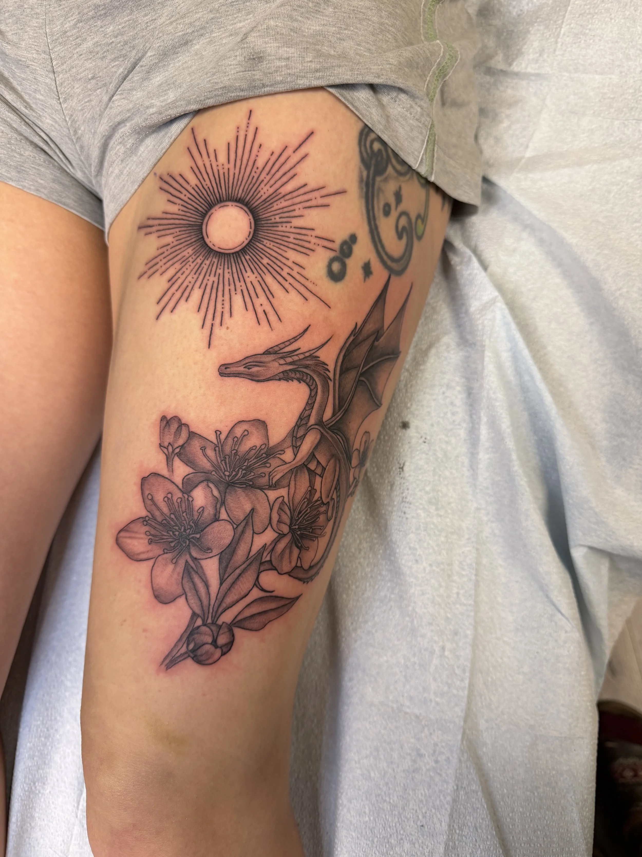 Tattoo on person's thigh features a sun with radiating lines, a moon shape inside the sun, a dragon with wings, and floral details.