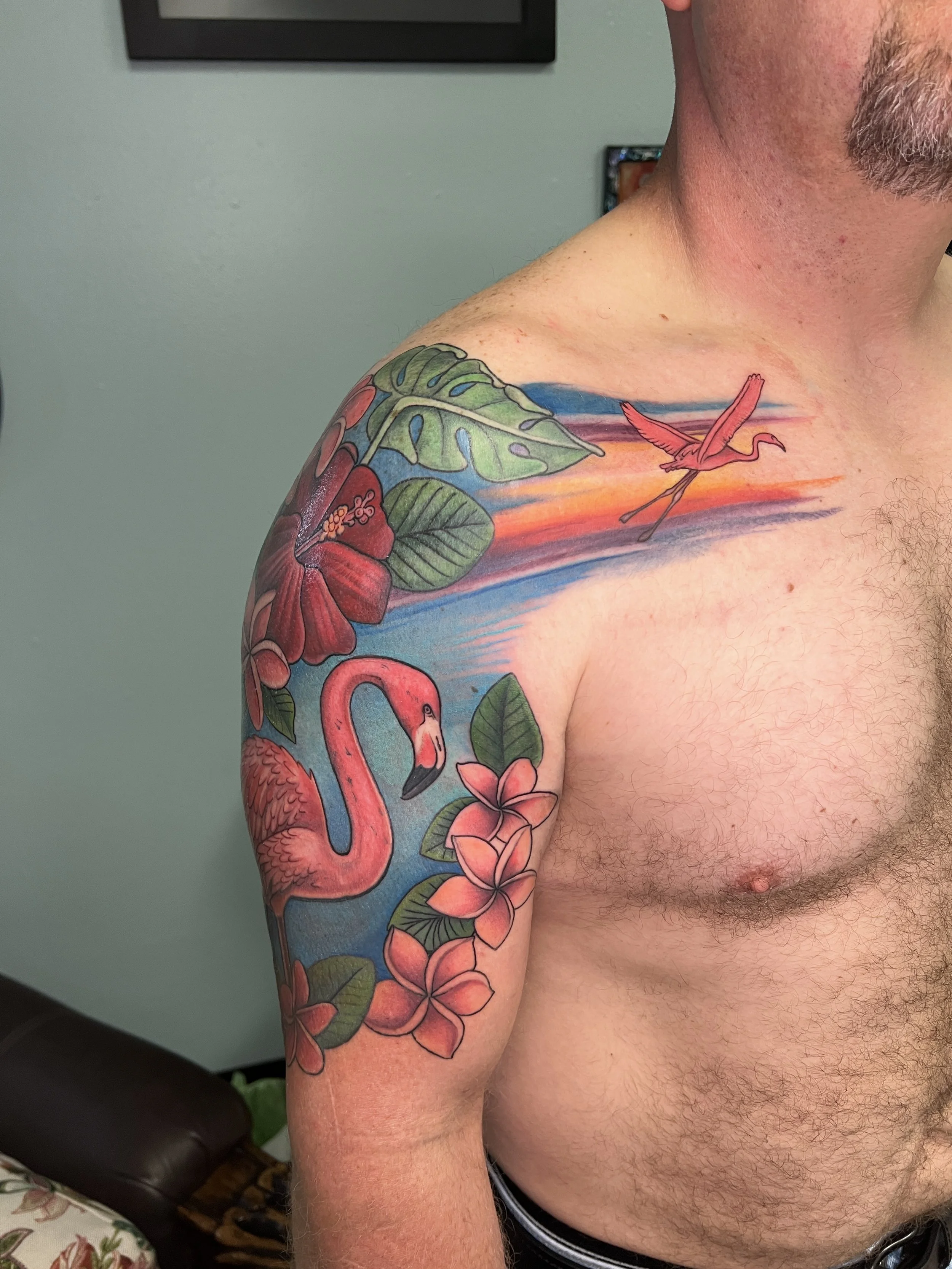 A person's upper arm and shoulder featuring a colorful tattoo of pink and red flowers, green leaves, and a flamingo with a background sunset with shades of orange, pink, and blue.