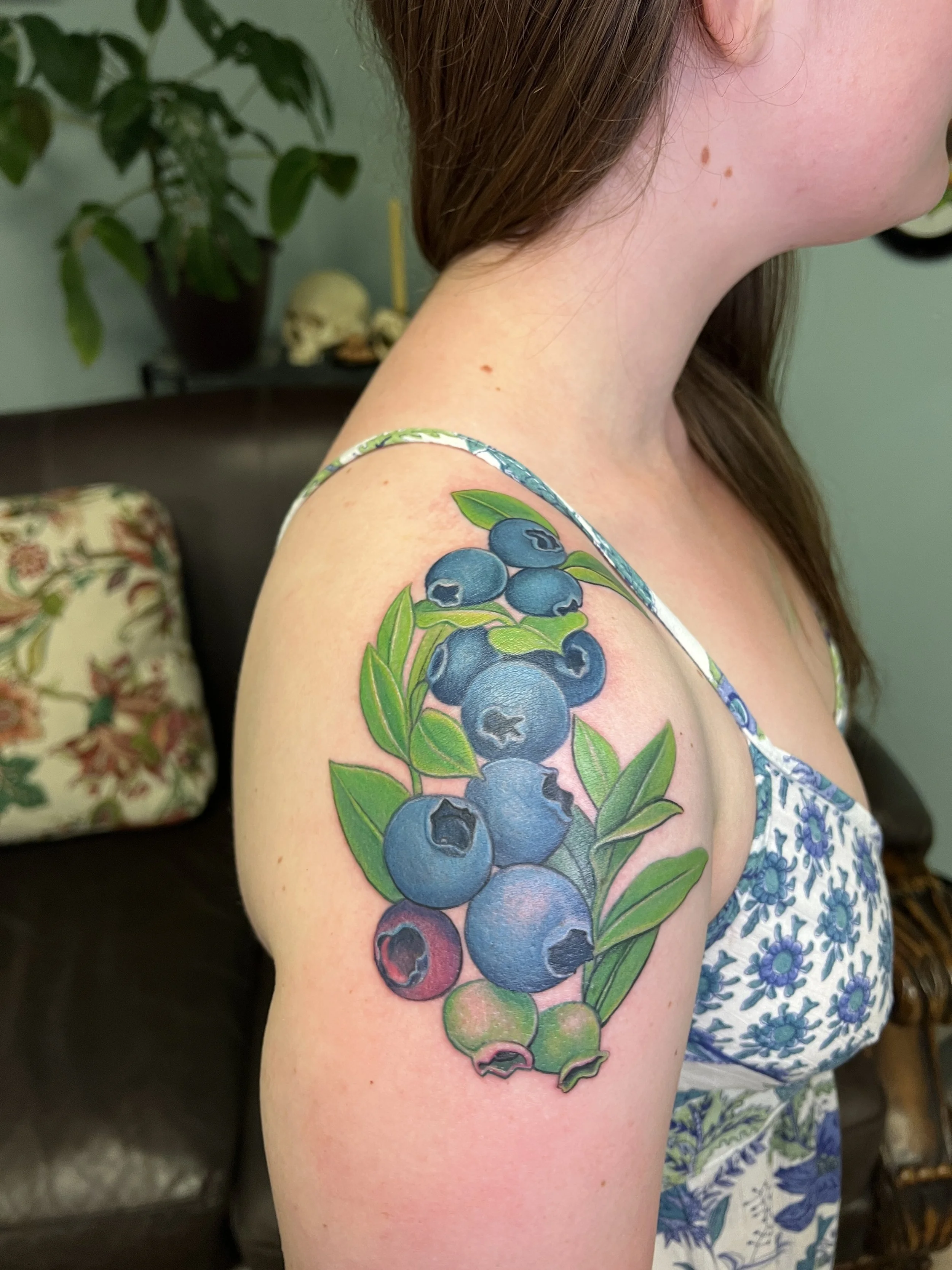 Close-up of a person's shoulder and neck with a colorful tattoo of blueberries and green leaves, wearing a blue and white floral dress, with part of the face and hair visible, indoor background with a potted plant, skull, and other decorative items.
