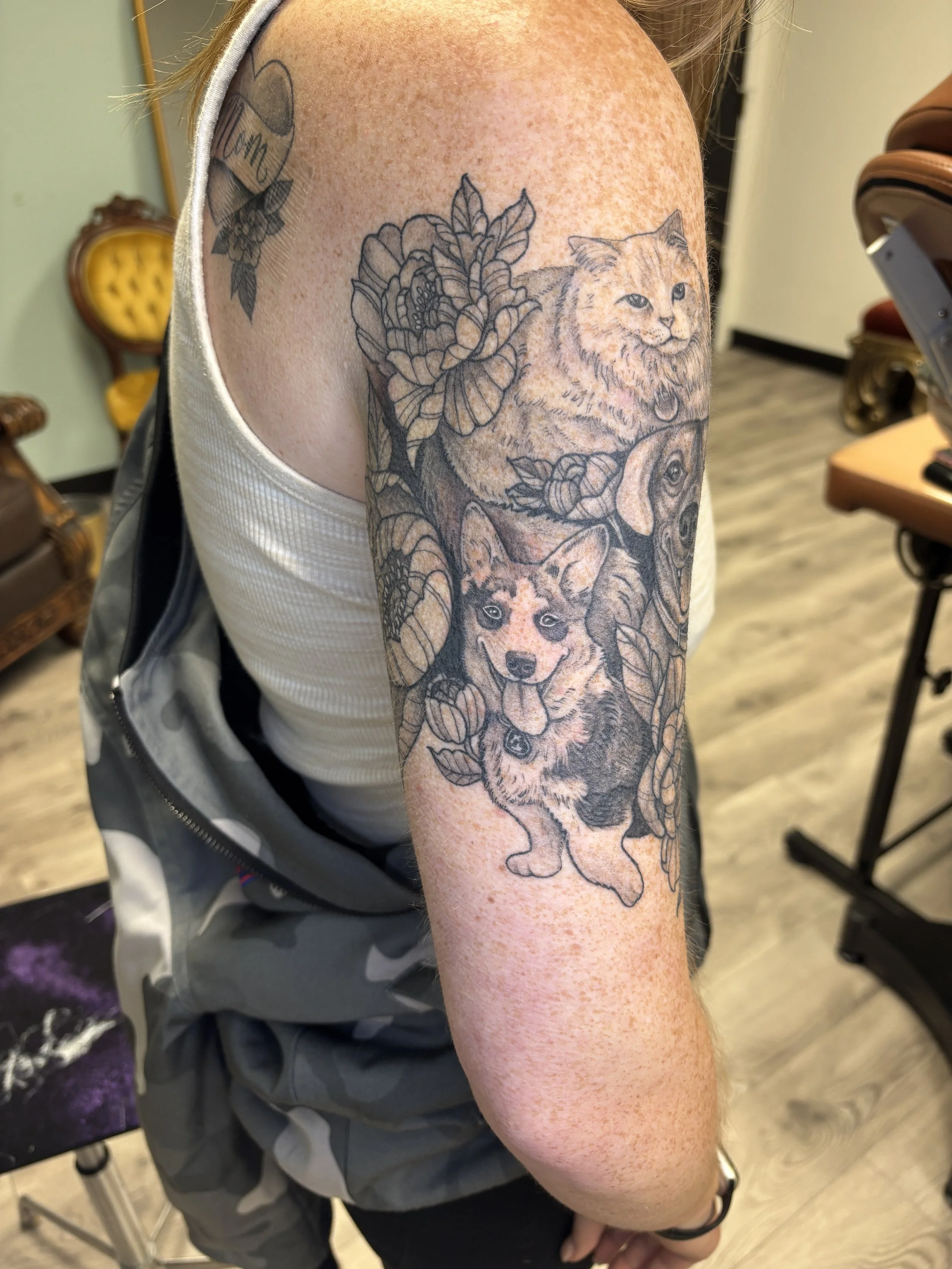 A person's upper arm with a detailed tattoo of a wolf surrounded by leaves and flowers, with the head of a cat within the design, and a heart tattoo with the word 'Mom' on the shoulder.