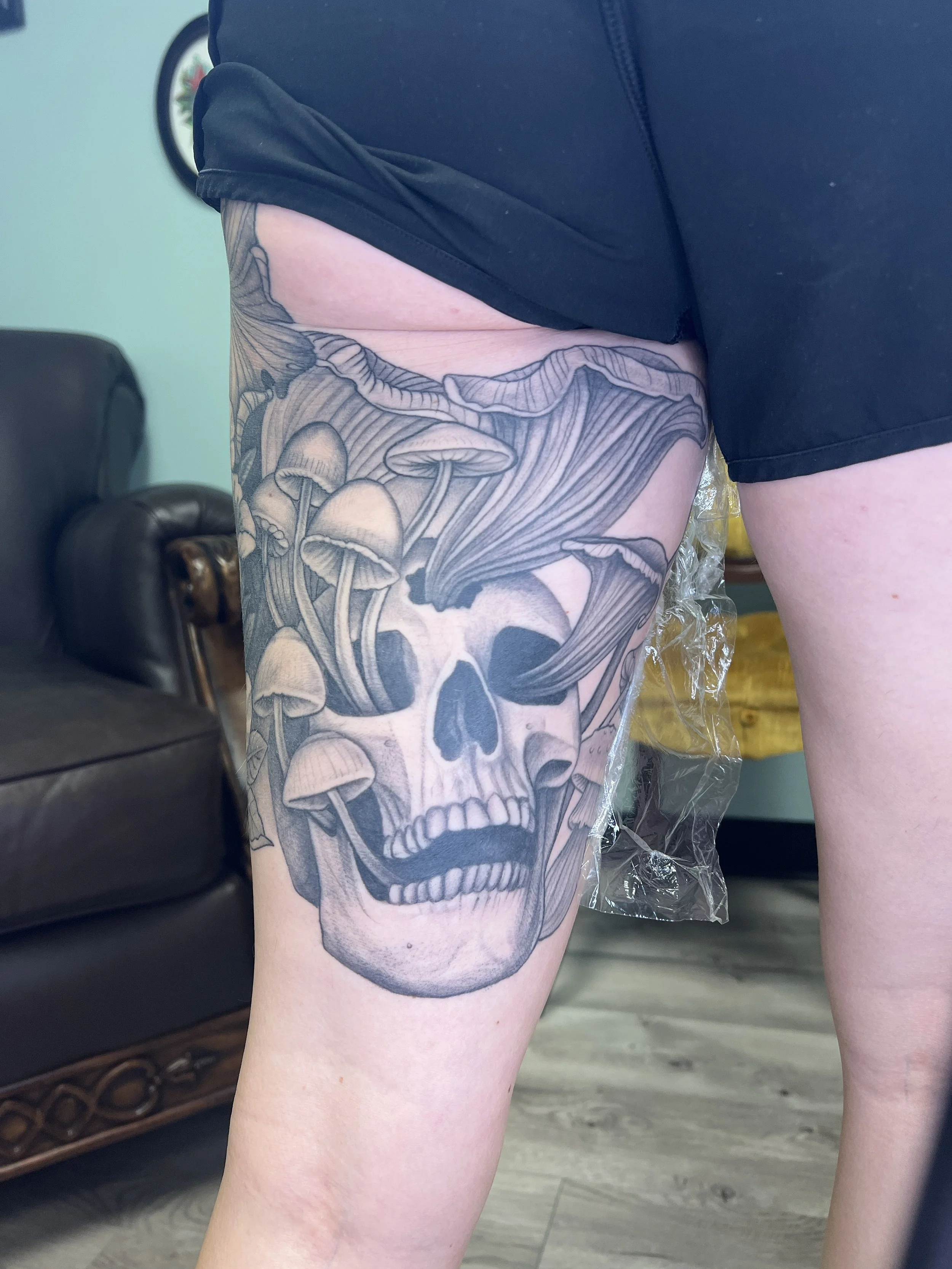 A person with a large black and white tattoo of a skull with mushrooms and forest elements on the back of their thigh