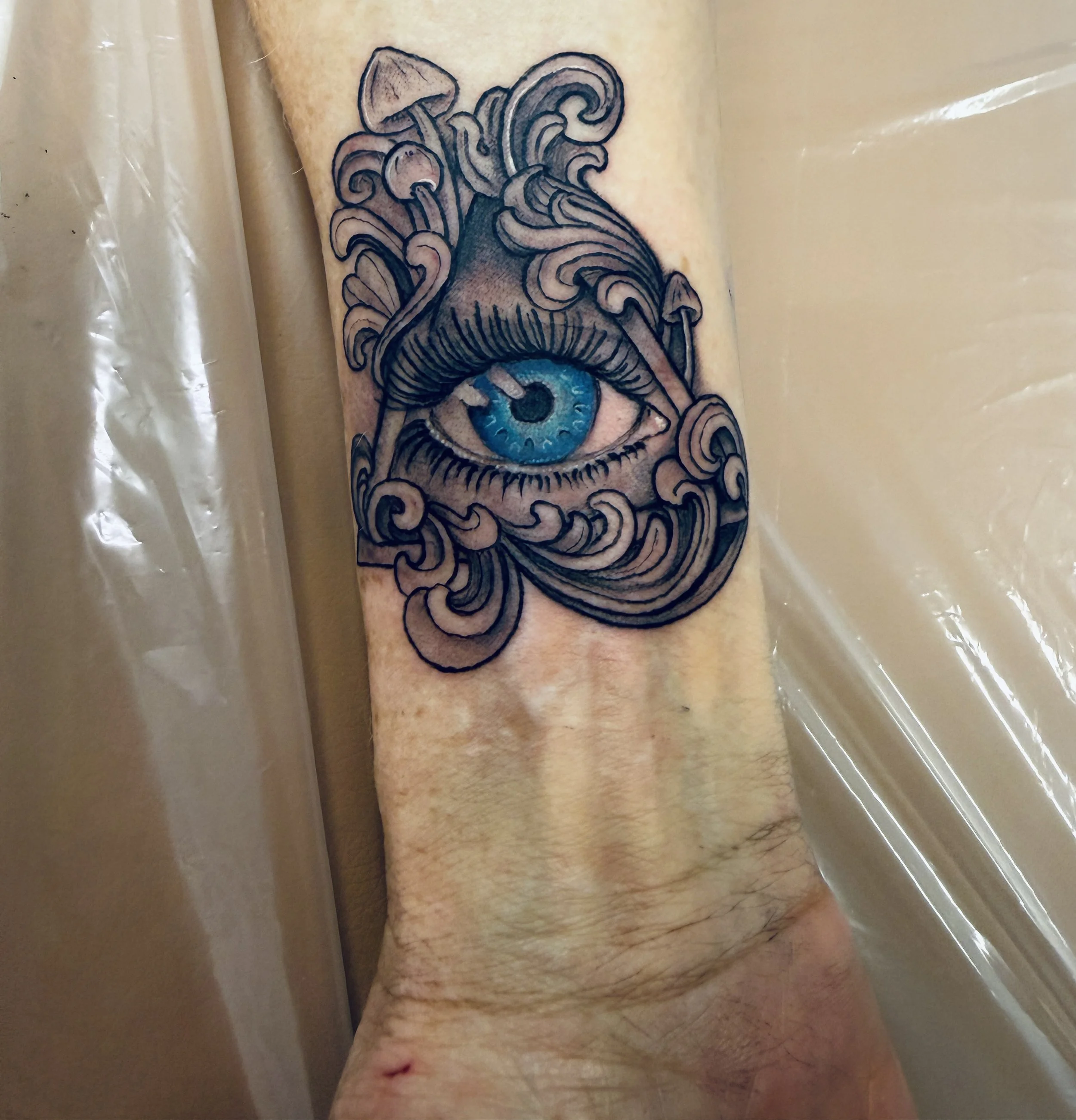 A tattoo of an eye with intricate swirling and mushroom tree designs around it, on a person's arm.