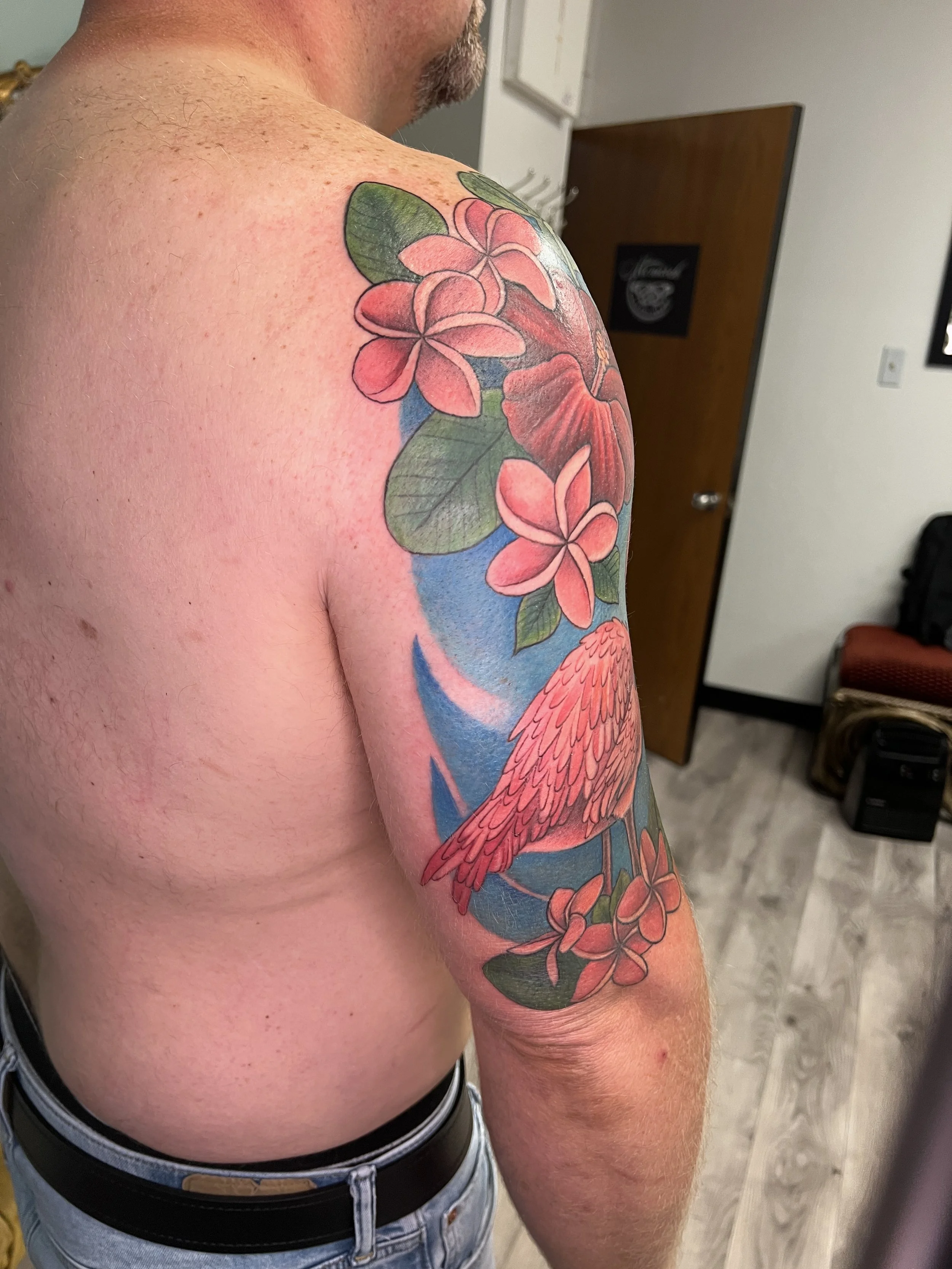 A person's upper arm and shoulder featuring a colorful tattoo of pink and red flowers, green leaves, and a flamingo