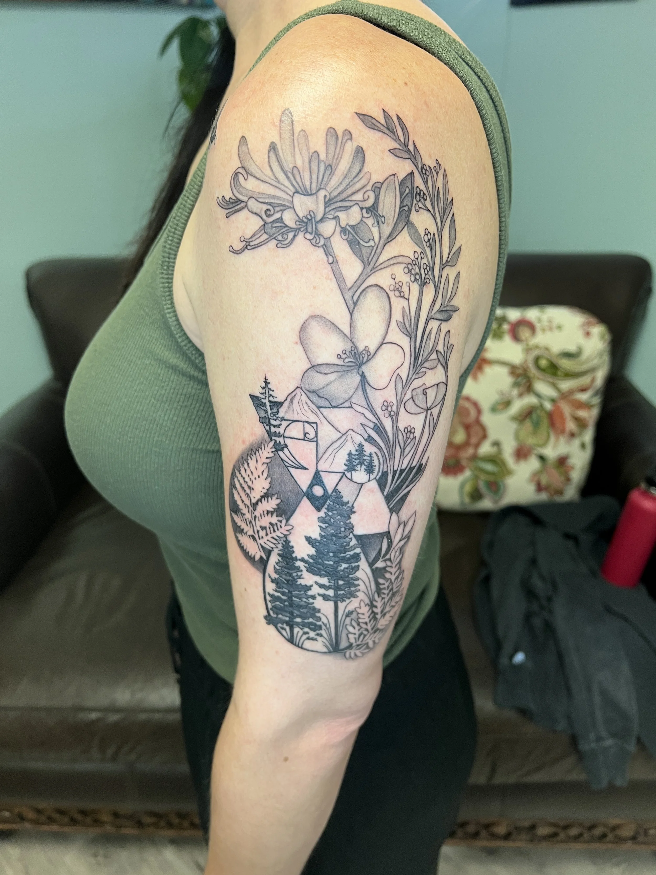 A person with a black and gray mountain and forest tattoo on their upper arm and shoulder, wearing a green sleeveless top.