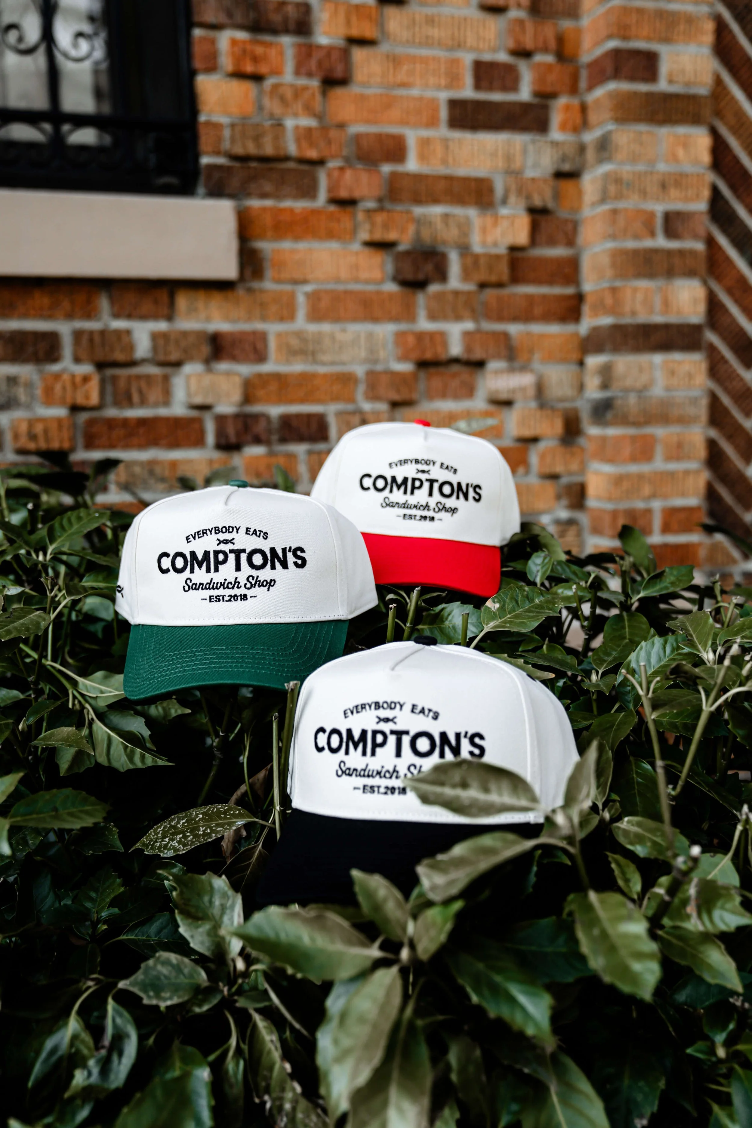 Several white and colored baseball caps with the logo for Compton's Sandwich Shop are placed among green leafy plants in front of a brick building with a window and black iron bars.