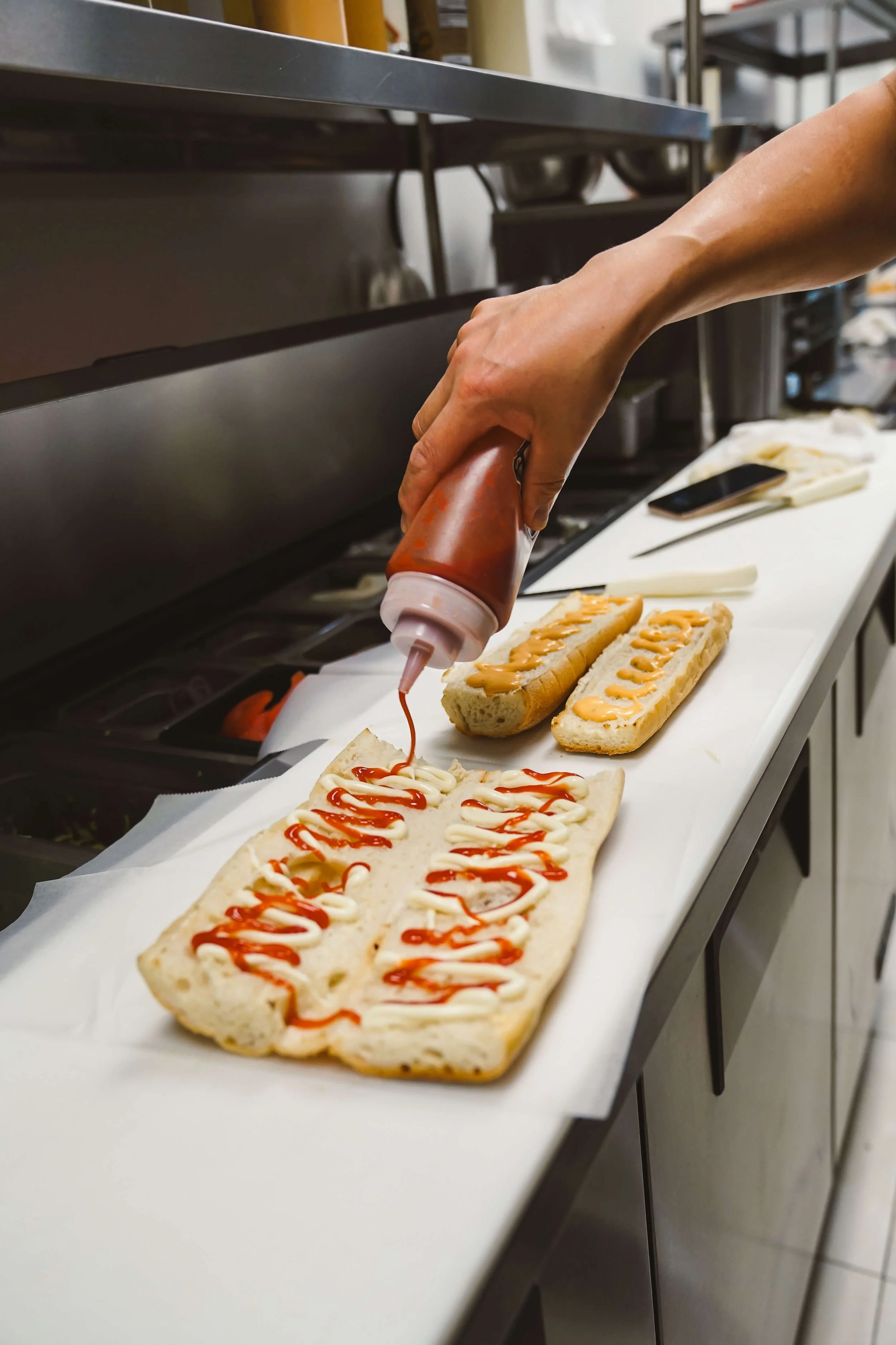 Person squeezing ketchup onto hot dog sandwiches with mayonnaise and mustard on a white papered surface in a kitchen or food prep area.