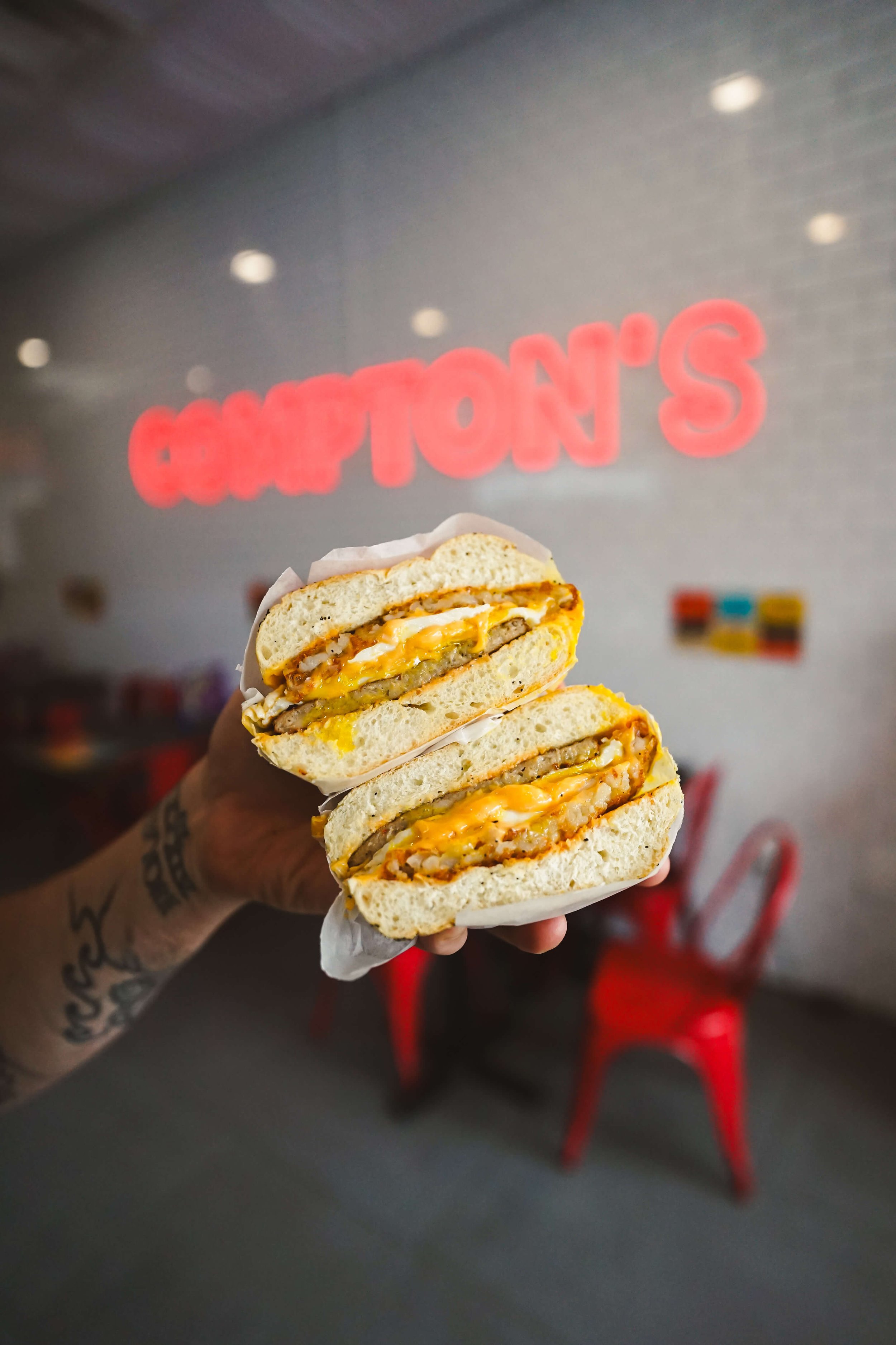 Person holding a half-eaten breakfast sandwich with egg, sausage, and cheese in front of a wall with a red and white sign for a restaurant or sandwich shop.