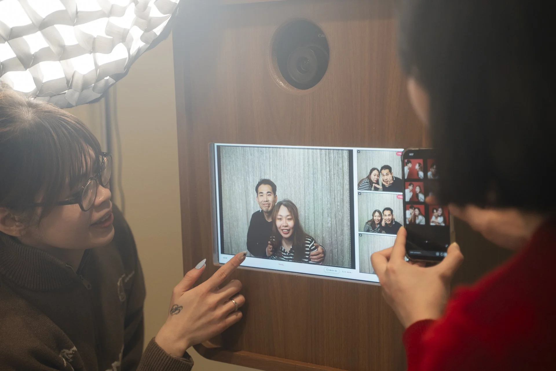 Two women take a selfie together inside a photo booth. They are smiling and appear happy. One woman has glasses and a tattoo on her finger. A person next to them holds a smartphone, capturing the photo. There is a computer screen showing the photo frame of the two women, and a camera is mounted on the wall above the screen.