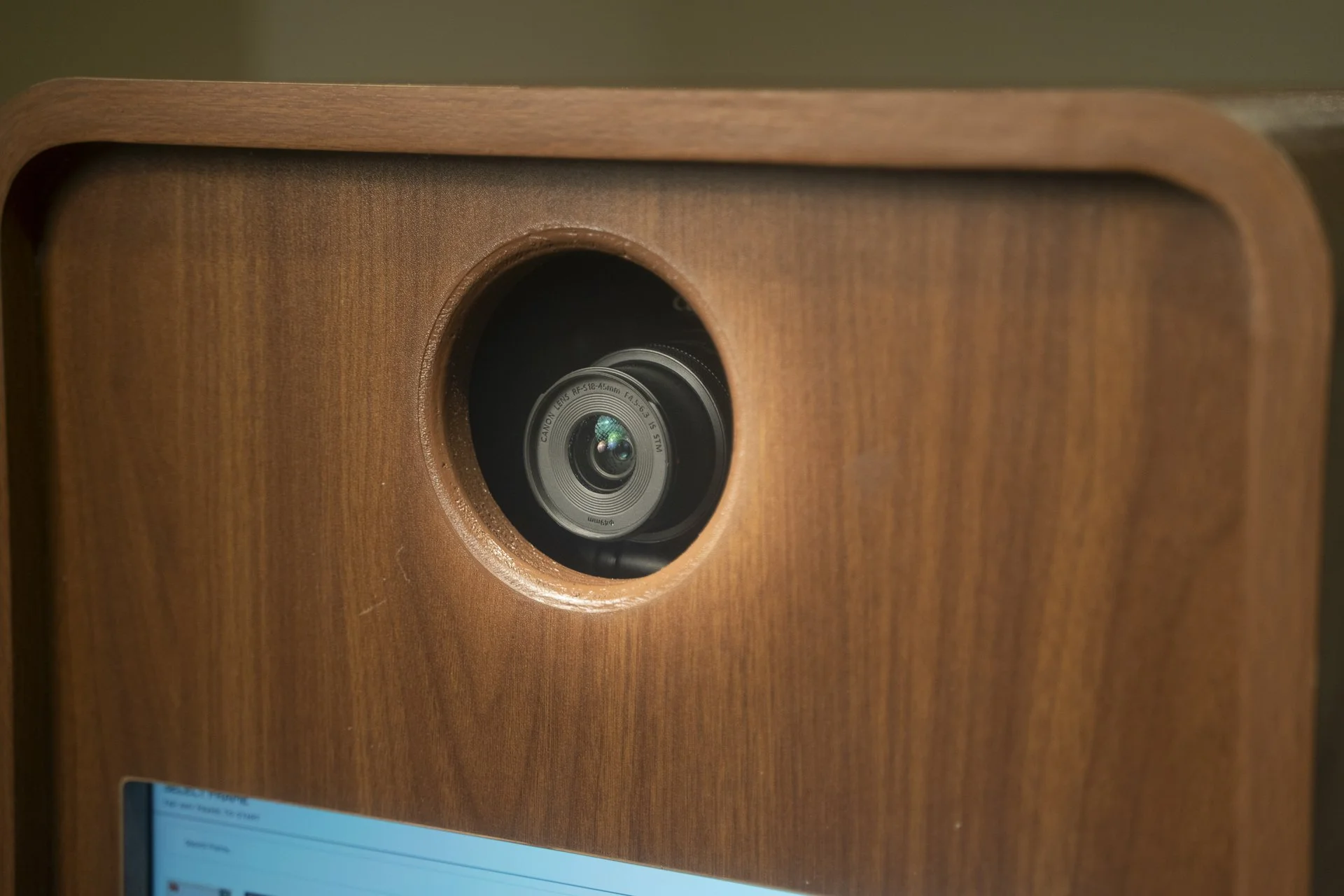 Close-up of a wooden photo booth with a visible lens, part of a larger electronic device, with a blue screen visible at the bottom.