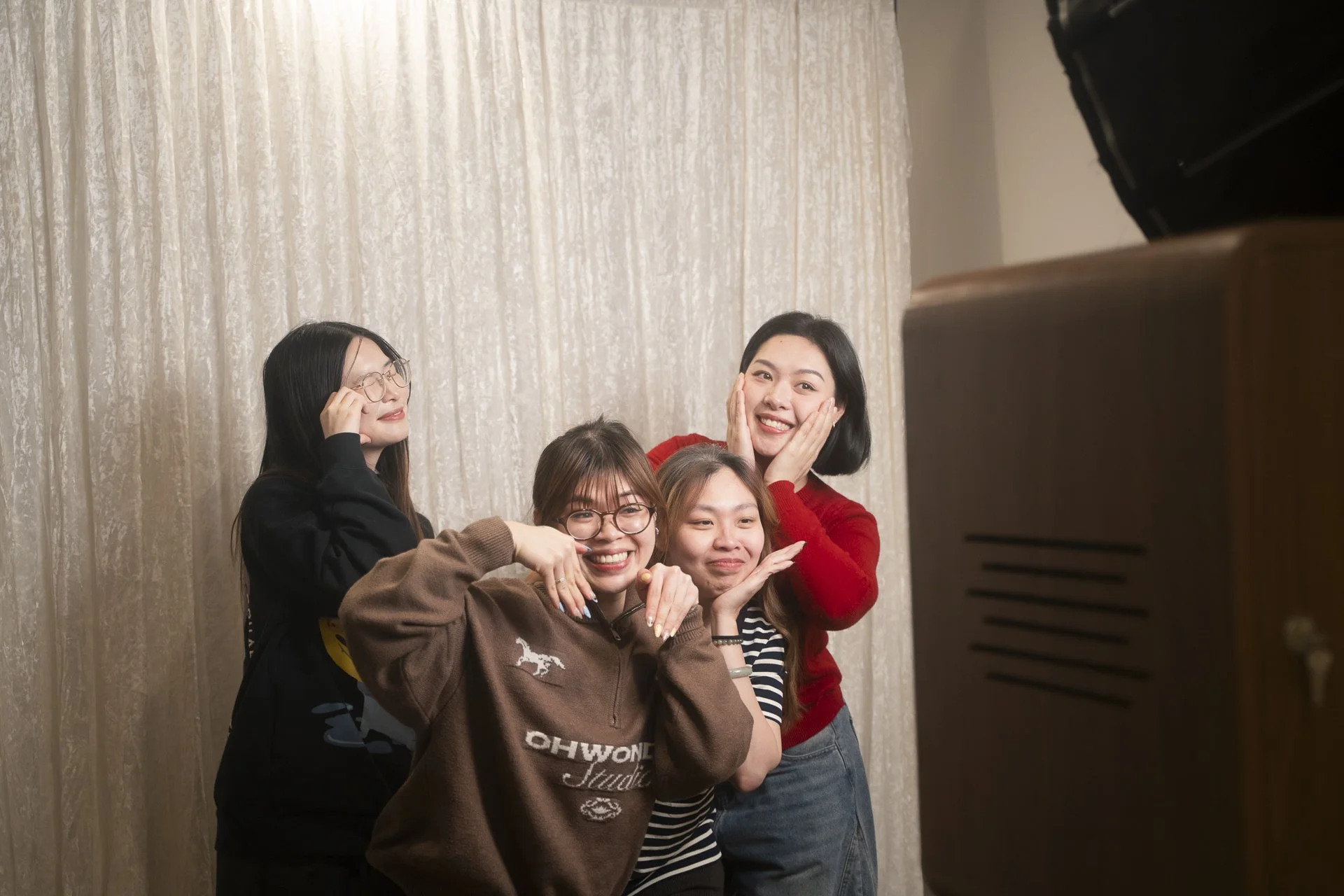 A group of five young women smiling and posing for a photo in front of beige curtains, some with glasses, casually dressed.