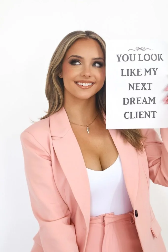A young woman with blonde hair wearing a pink suit and white top, holding a sign that says 'You look like my next dream client'.