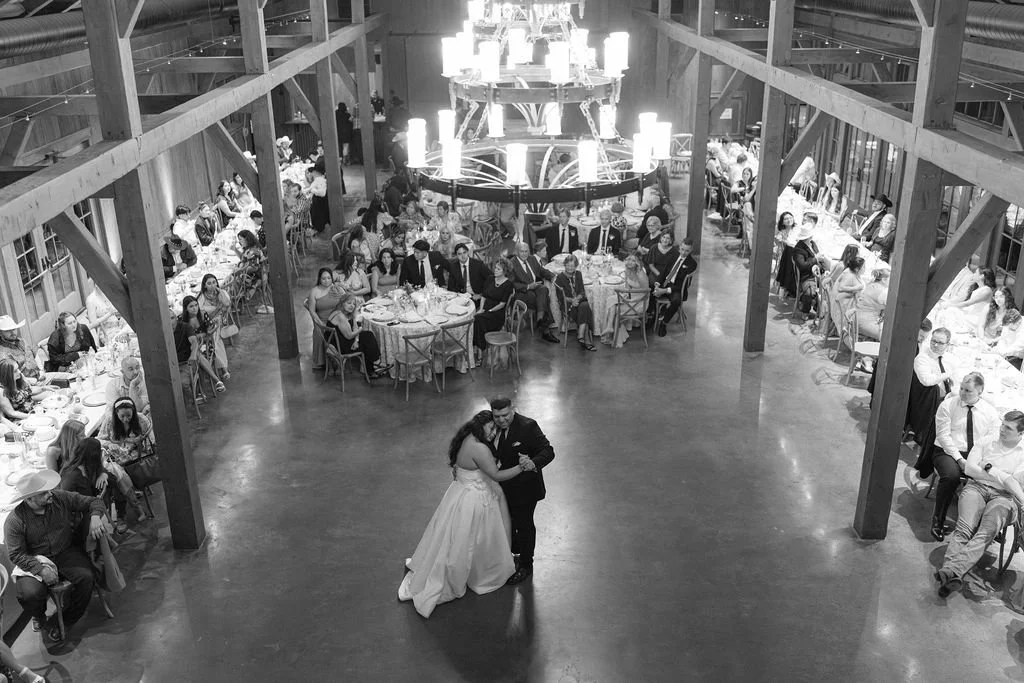 A bride and groom dancing in a rustic barn wedding reception with guests seated at round tables around them and a large chandelier overhead.