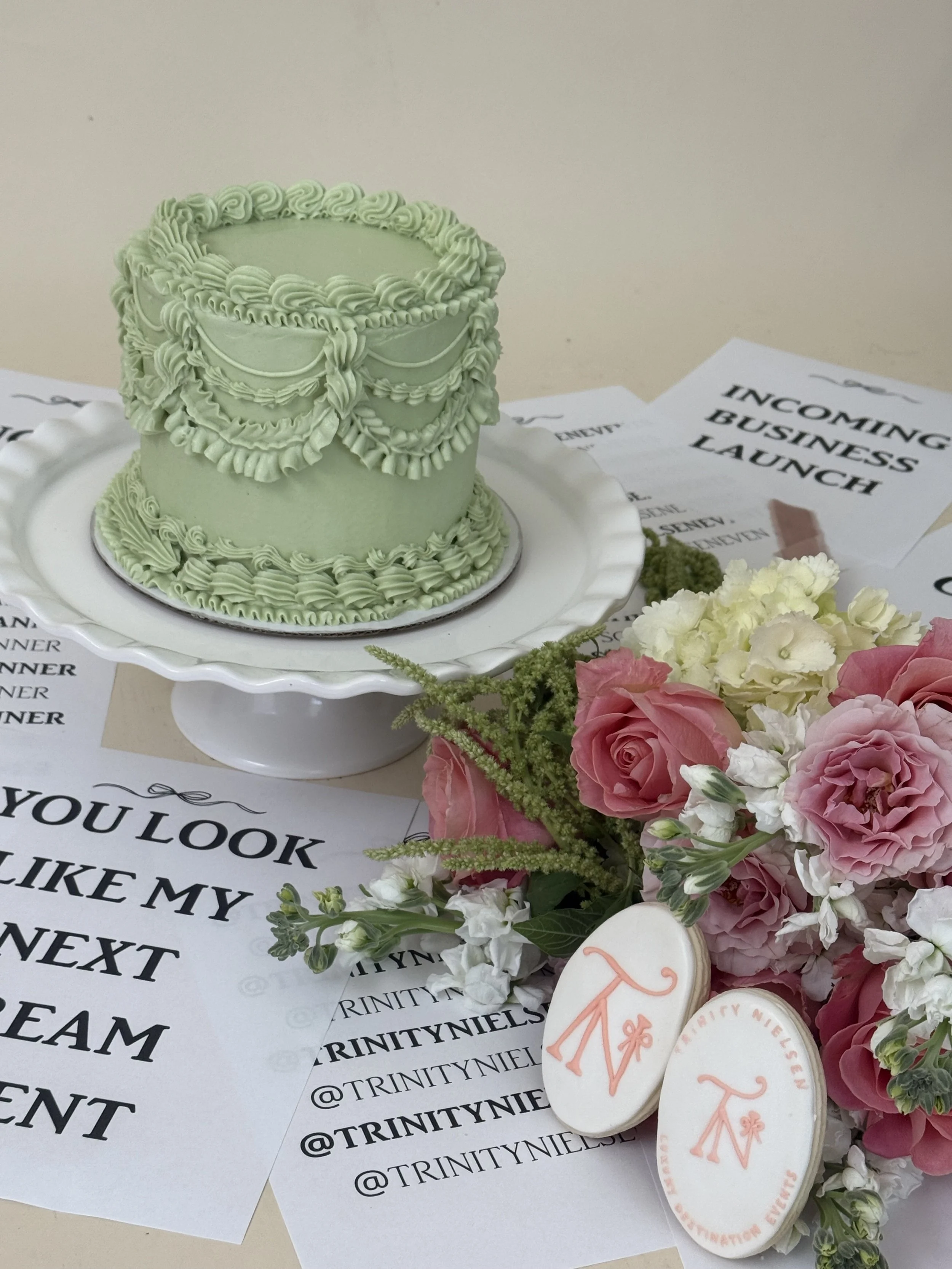 A light green decorated cake with piped frosting and a floral border on top, placed on a white cake stand. Surrounding the cake are pink and white flowers, along with some of the printed papers that read 'INCOMING BUSINESS LAUNCH' and social media handle '@TRINITYNIELS'.