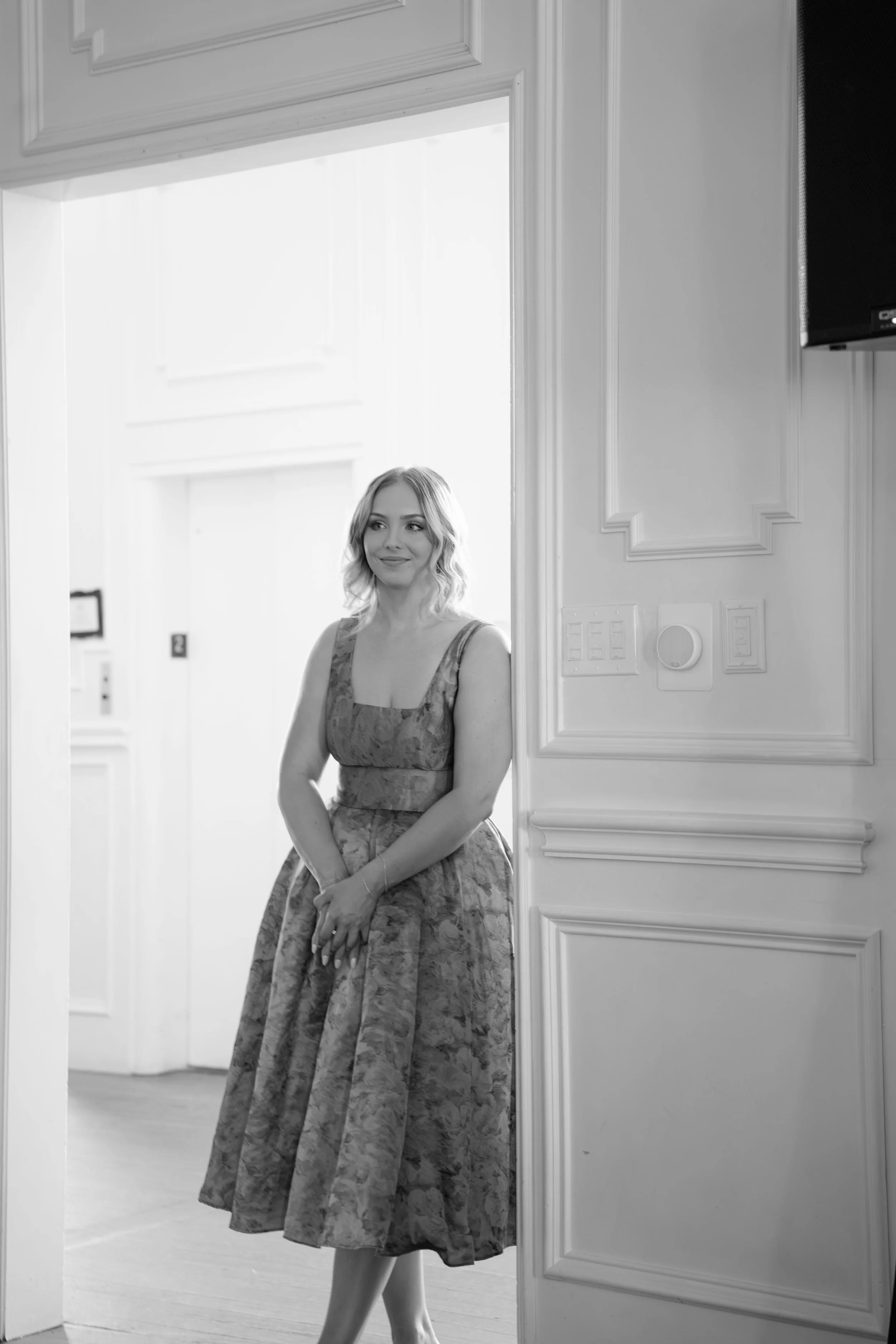 A young woman with blonde hair wearing a floral dress standing near a door in a bright room.