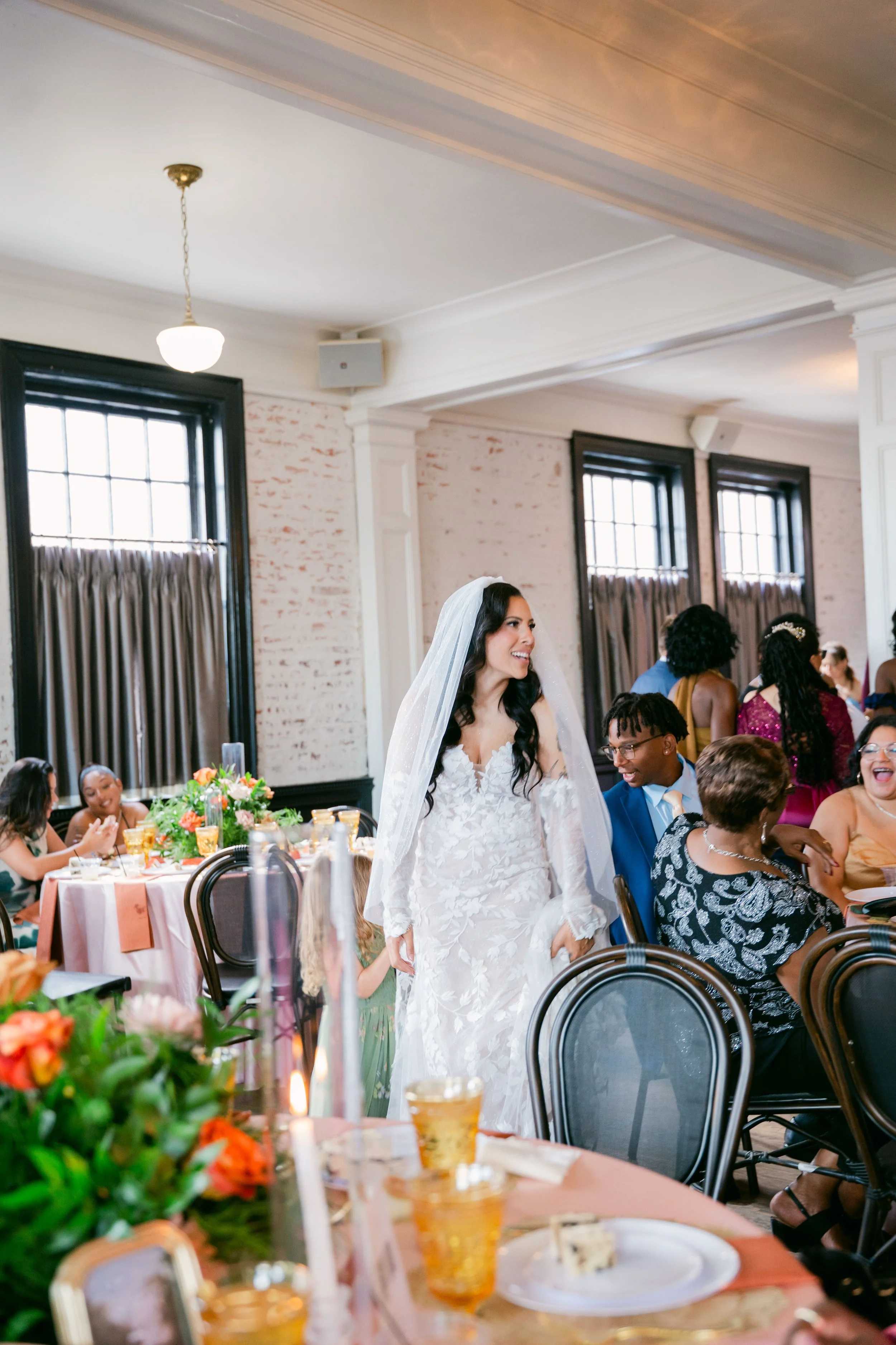 A bride in a white wedding gown with a veil smiling and holding hands with a young girl in a green dress at a wedding reception. Guests are seated at decorated tables with floral arrangements, and the room has high ceilings, large windows with curtains, and is filled with people enjoying the celebration.