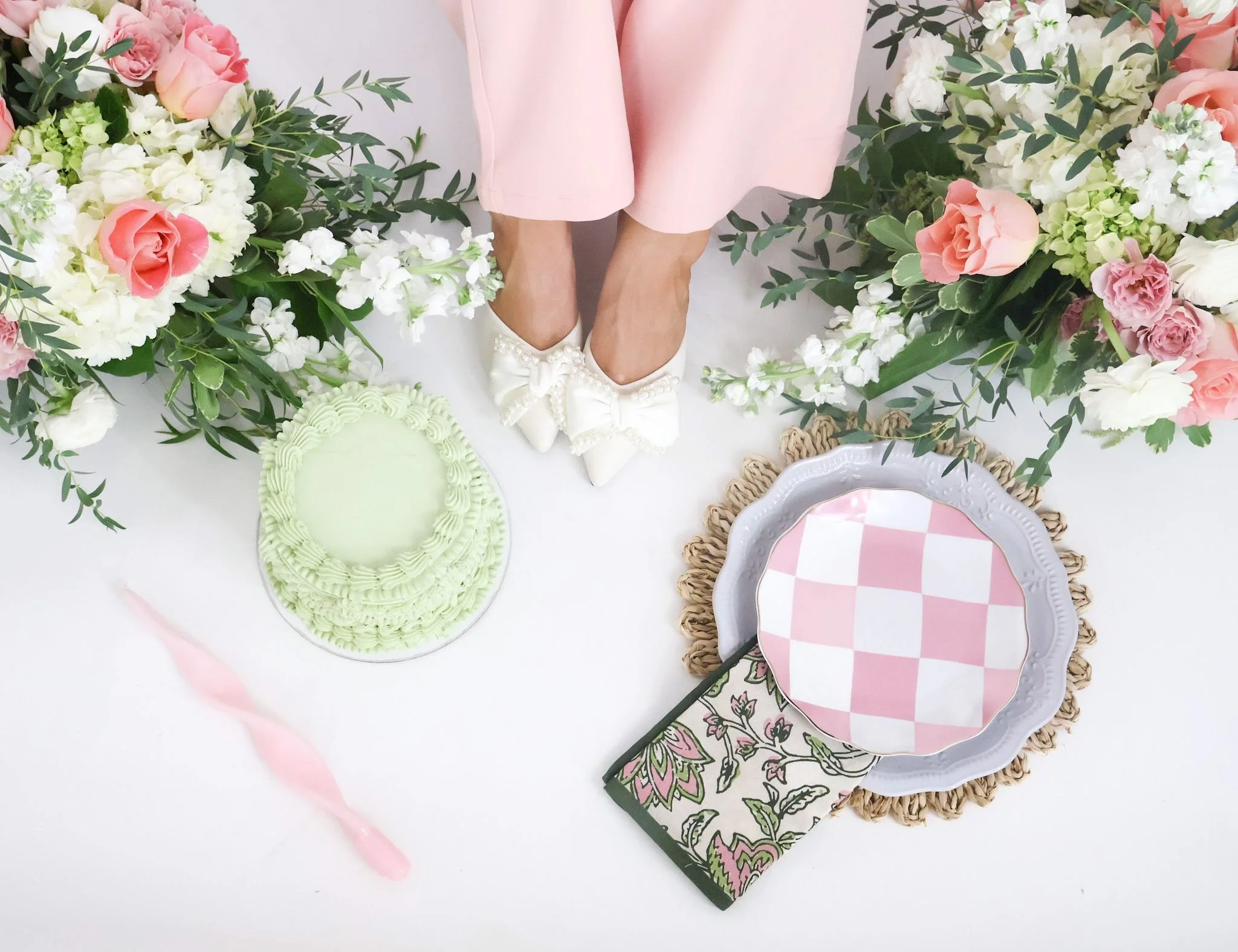 The image shows a pair of legs in pink pants, standing among floral arrangements with pink and white flowers. There are also pastel-colored plates and a decorative hand mirror on the table, suggesting a celebration or special occasion.