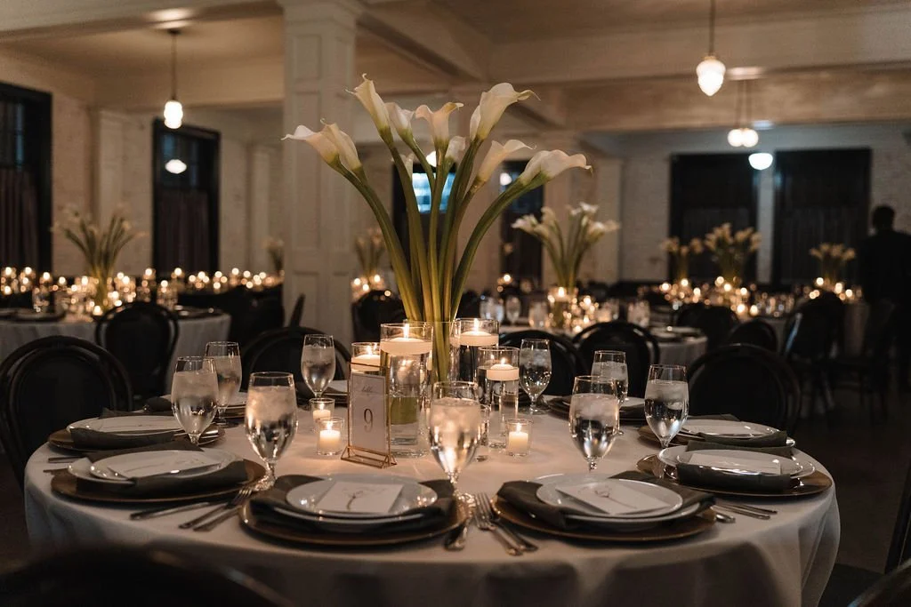 Elegant banquet table set with white tablecloth, floral centerpiece of white calla lilies, candles, glasses, plates, and silverware in a dimly lit event hall.