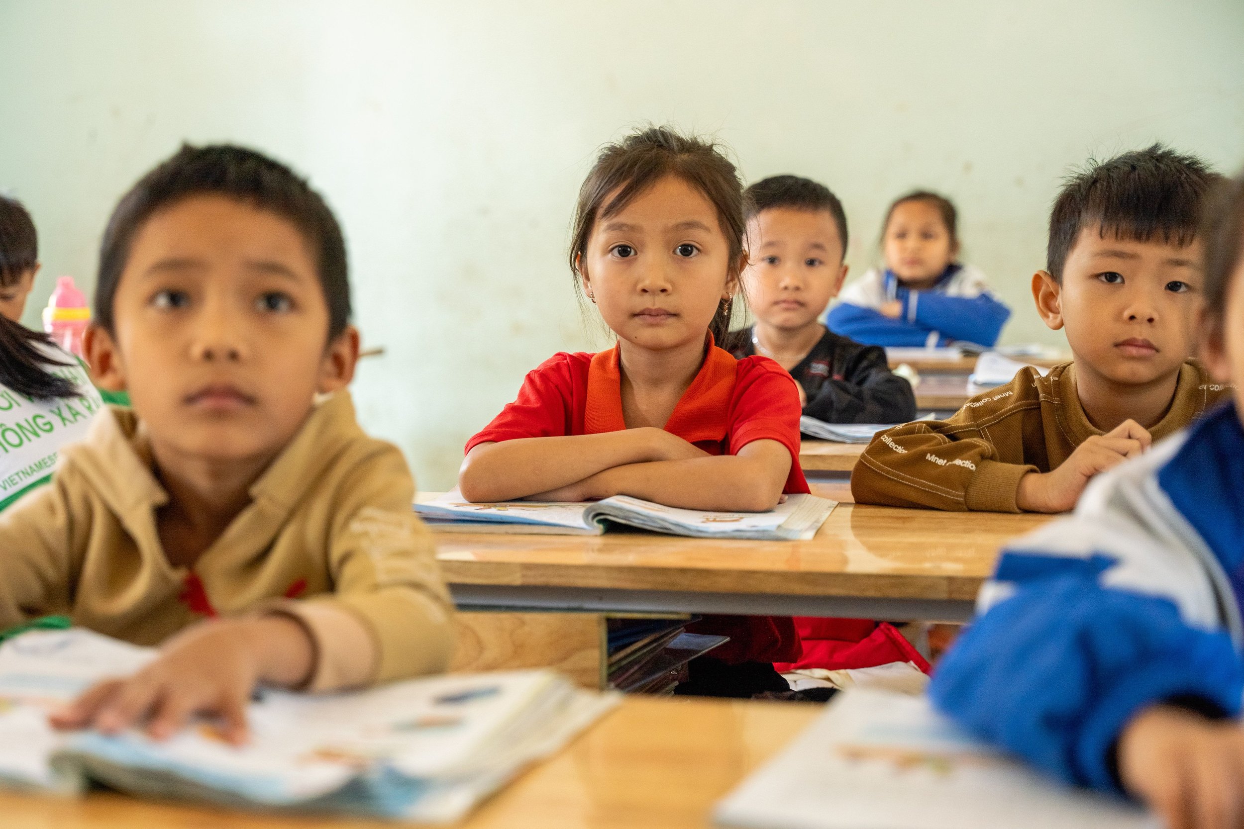 Young students seated at desks in a classroom, attentively listening to the teacher, with notebooks and school supplies in front of them.