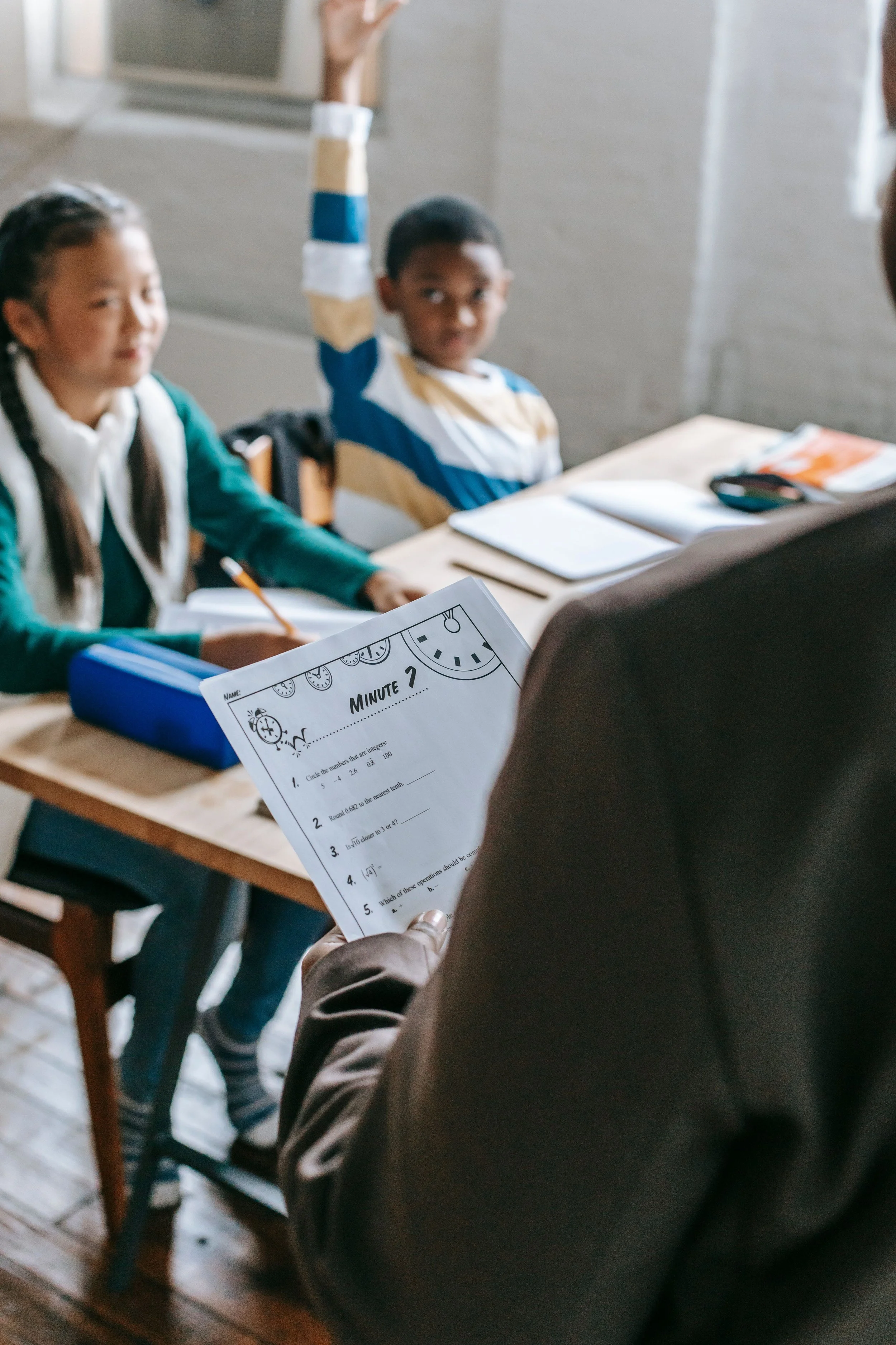 A classroom scene with students and a teacher or instructor. One student, a boy, is raising his hand. The teacher or instructor is holding a math worksheet, and there are open notebooks and a calculator on the desk. The students are seated at desks, and the classroom has a warm, well-lit atmosphere.