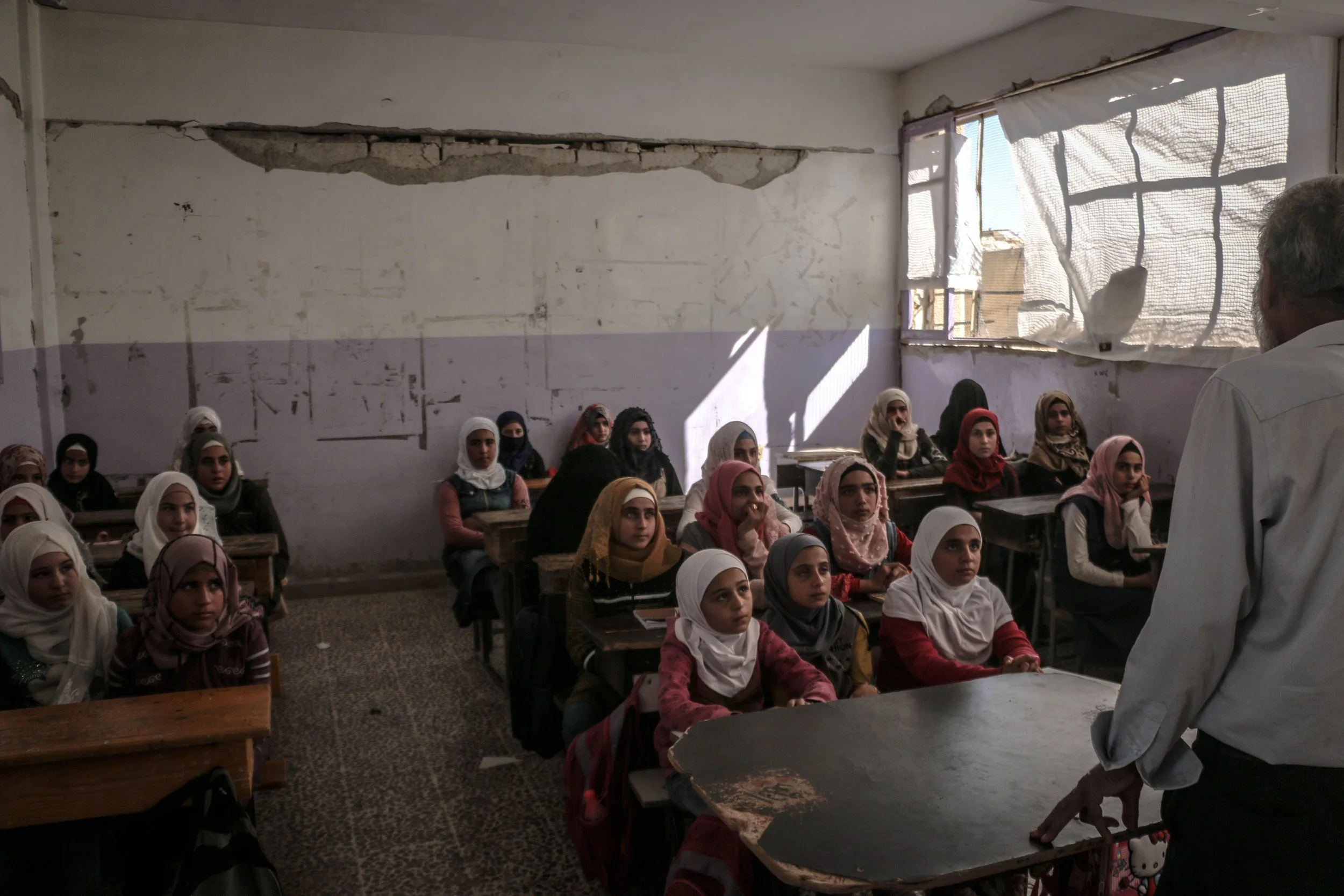 A classroom filled with young girls wearing headscarves, sitting at desks and listening to a male teacher standing in front of them. The classroom has a damaged wall and a torn window covering.