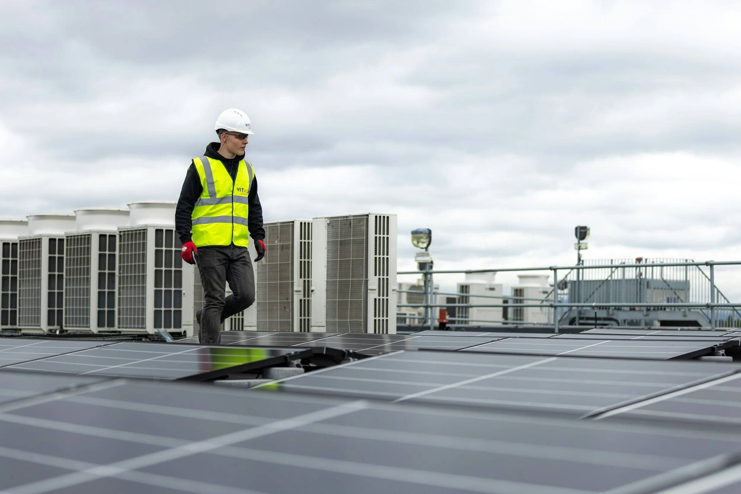 A person wearing a white hard hat, safety glasses, red gloves, a yellow safety vest, black hoodie, and dark pants walking on a rooftop with solar panels and HVAC units, under a cloudy sky.
