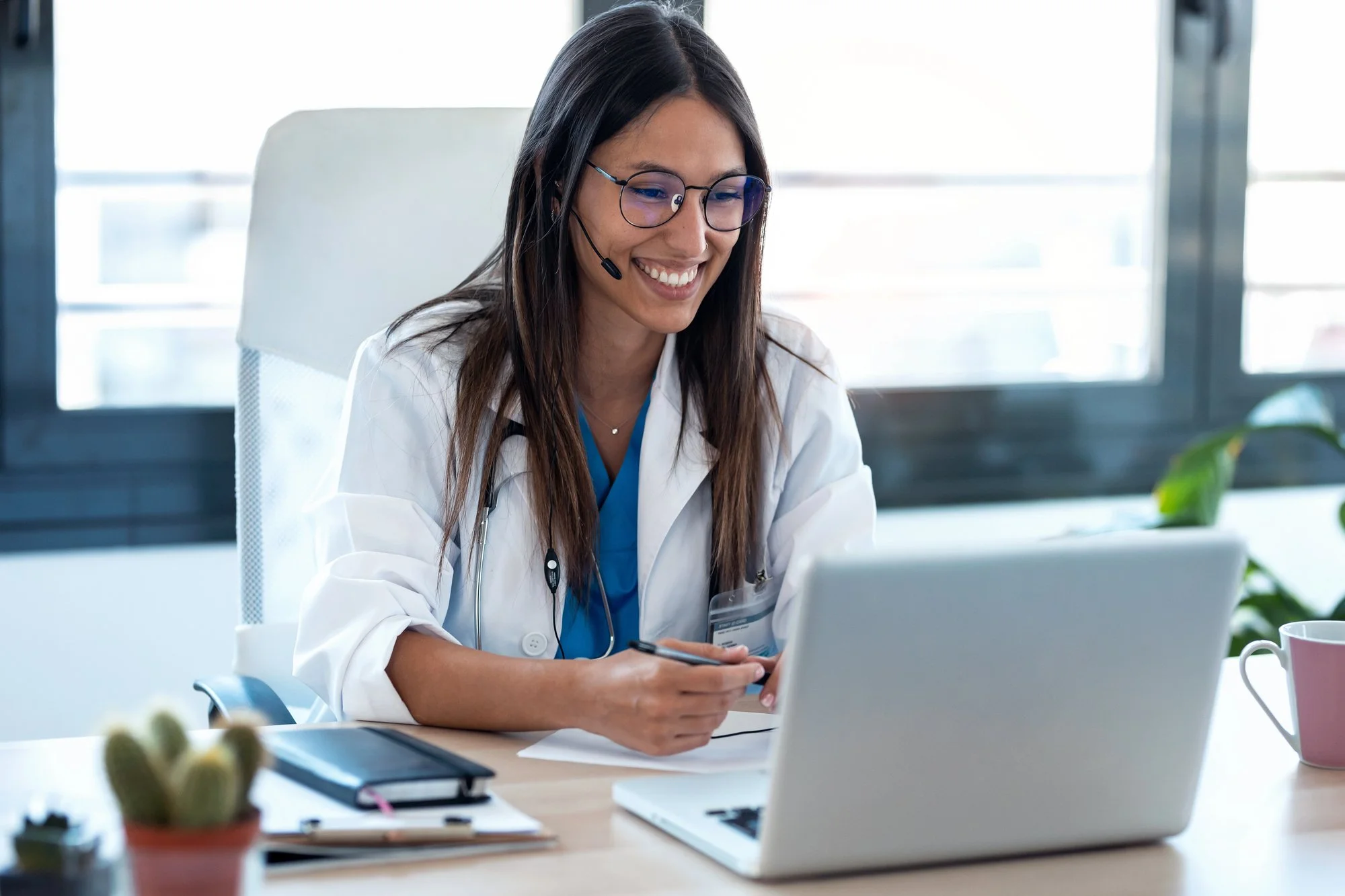 A smiling female healthcare worker with long brown hair, glasses, and a headset, sitting at a desk using a tablet in an office with large windows.