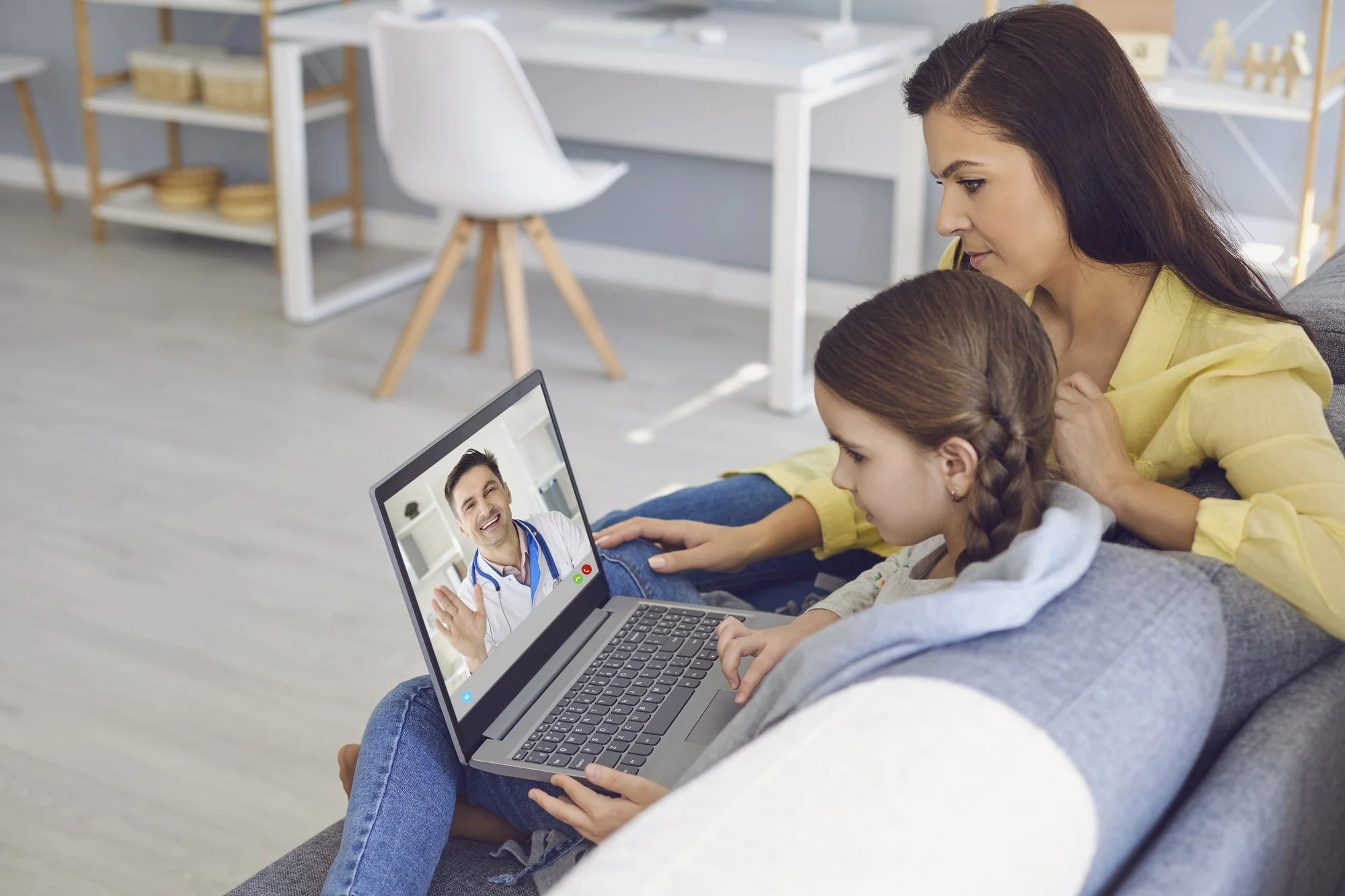 A woman and a girl on a gray couch during a video call with a smiling male doctor on a laptop, in a modern, well-lit living room.