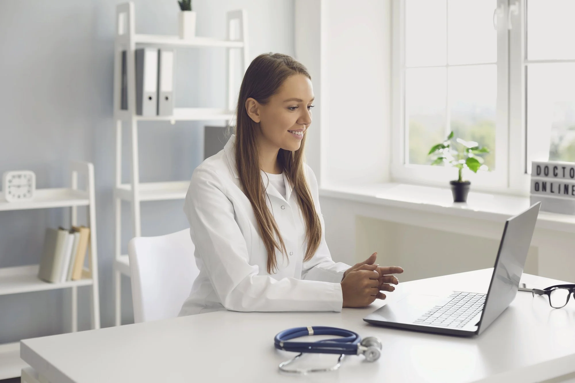 A female doctor in a white coat sitting at a desk with a laptop, stethoscope, and computer in a bright, modern office with large windows and a plant.