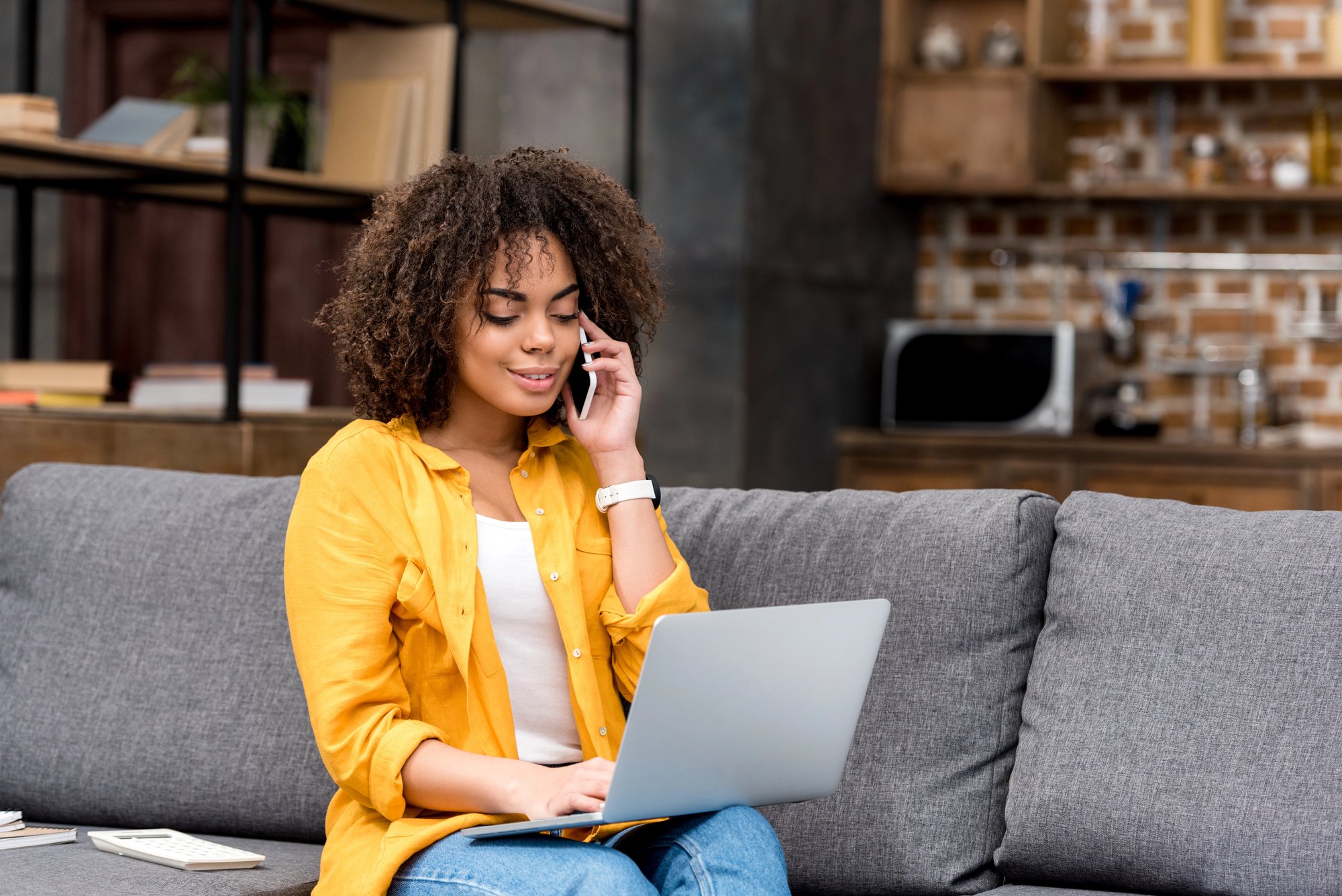 Young woman with curly hair wearing a yellow shirt, talking on the phone and using a laptop, sitting on a gray sofa in a modern living room with shelves and brick wall in the background.