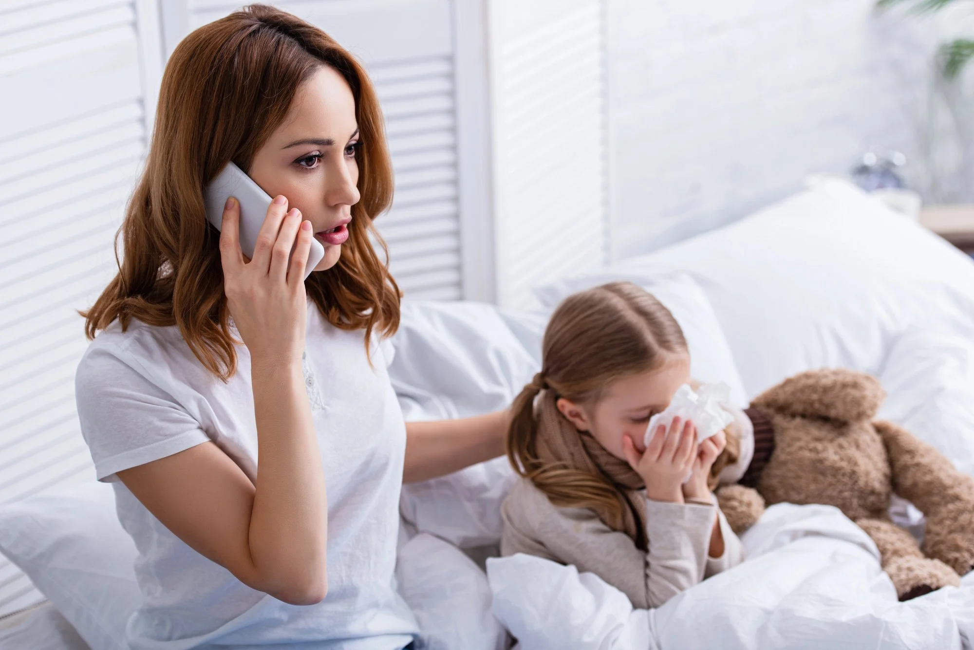 A woman with red hair on the phone sitting on a bed with two children, one with red hair and the other with brown hair, both holding tissues and tissues in hand, in a bright room.