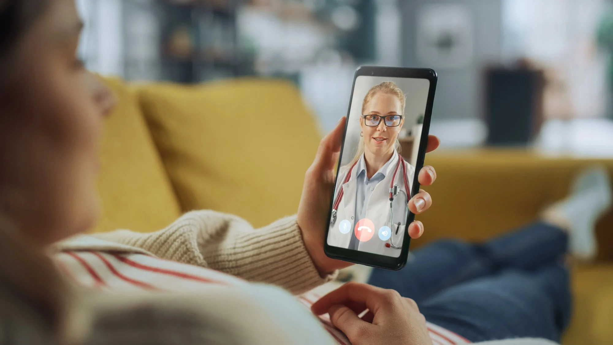 Person holding a smartphone during a video call with a female doctor wearing glasses and a white coat with a stethoscope, sitting on a yellow couch in a room with blurred background.