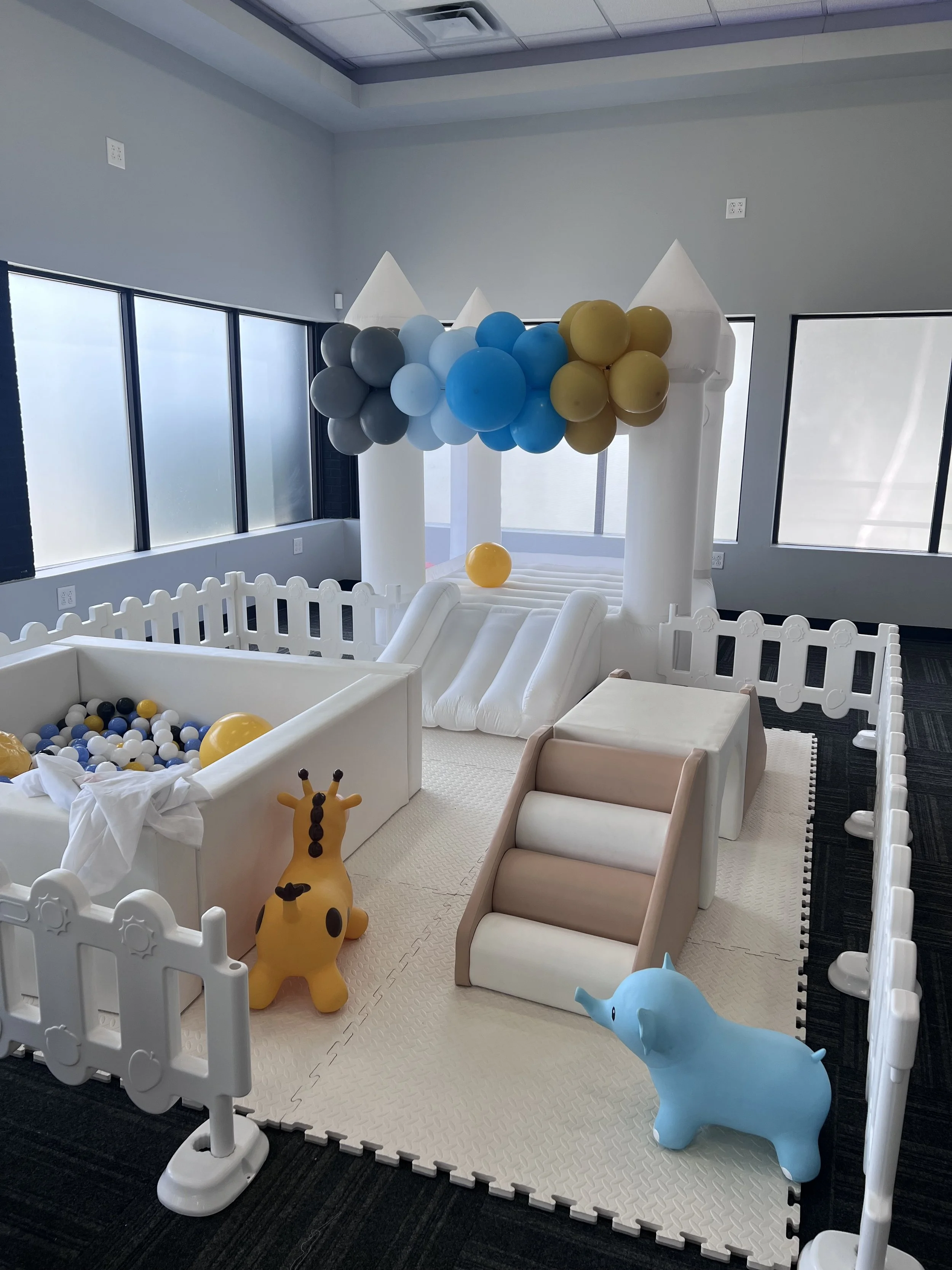 Indoor play area with white inflatable castle featuring small slide, surrounded by plastic animal toys including a giraffe and an elephant, enclosed by a white picket fence, with balloon decorations in blue, gray, and yellow hanging from the castle.