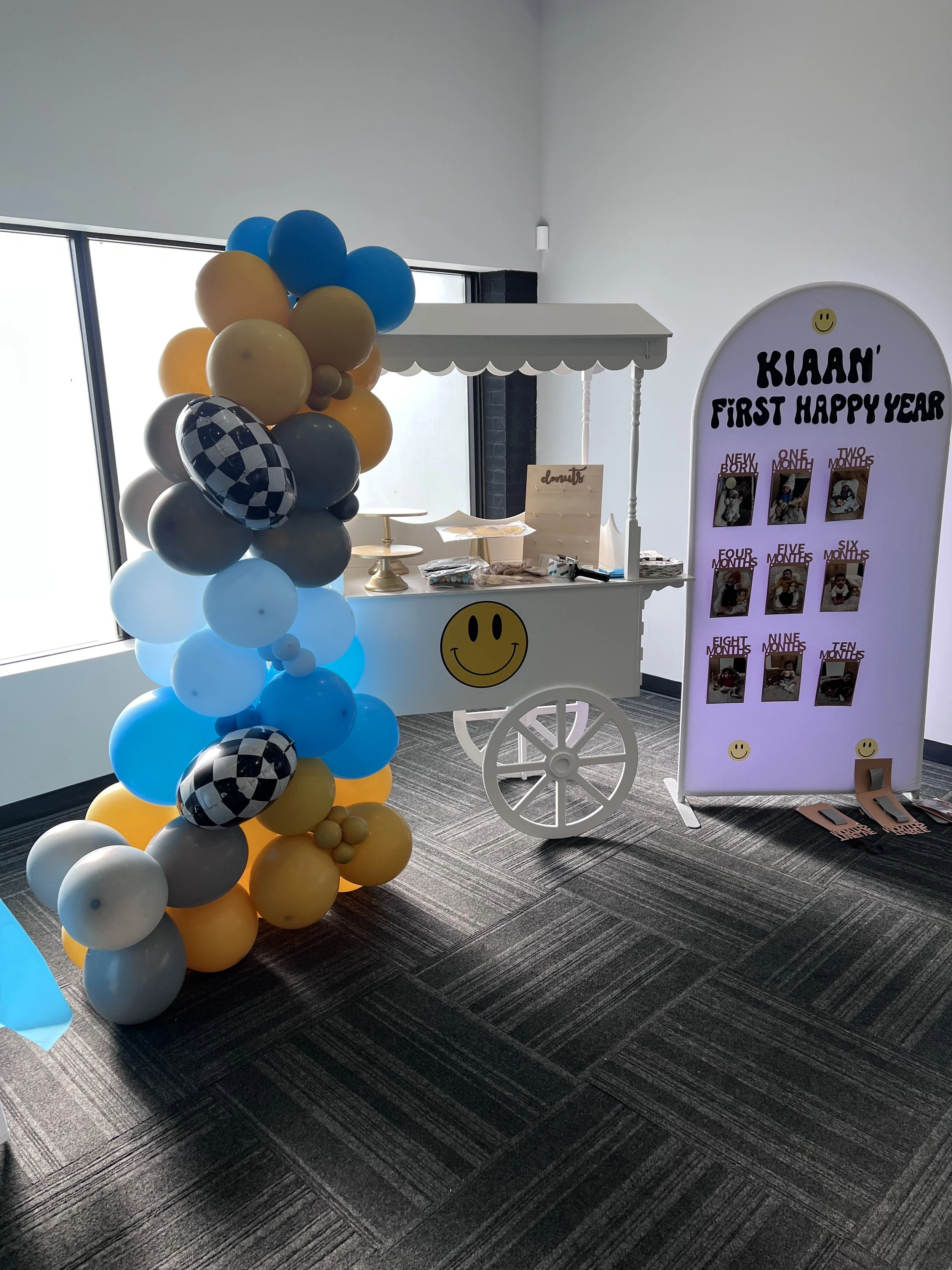 Decorative display for a baby's first year celebration with a balloon arrangement of blue, yellow, gray, and checkered balloons, a small vendor cart with a smiley face, and a white board with photos of the baby at different months.