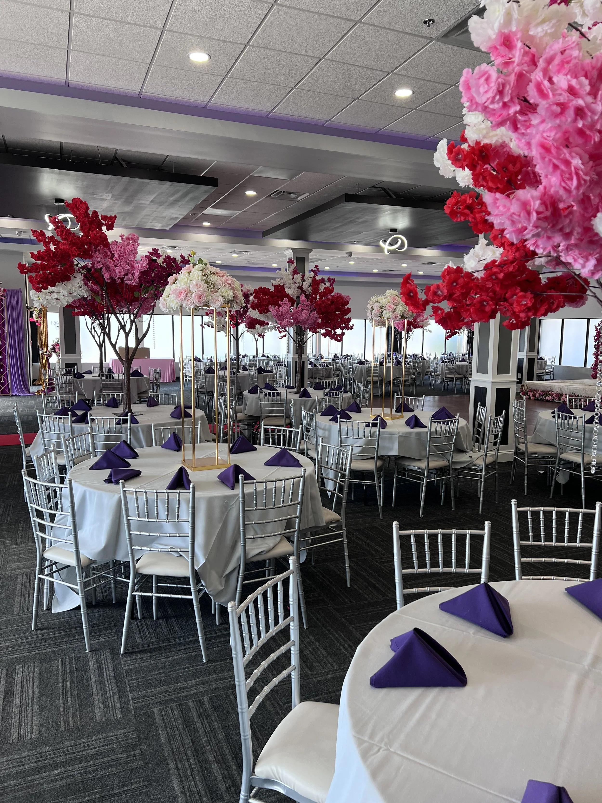 A decorated banquet room with round tables covered with white tablecloths, each with purple napkins folded on top. Tall floral centerpieces with pink, red, and white flowers are set on golden stands. Silver chairs surround the tables. Large windows line the back wall, allowing natural light into the room. Overhead lighting and decorative ceiling features are also visible.