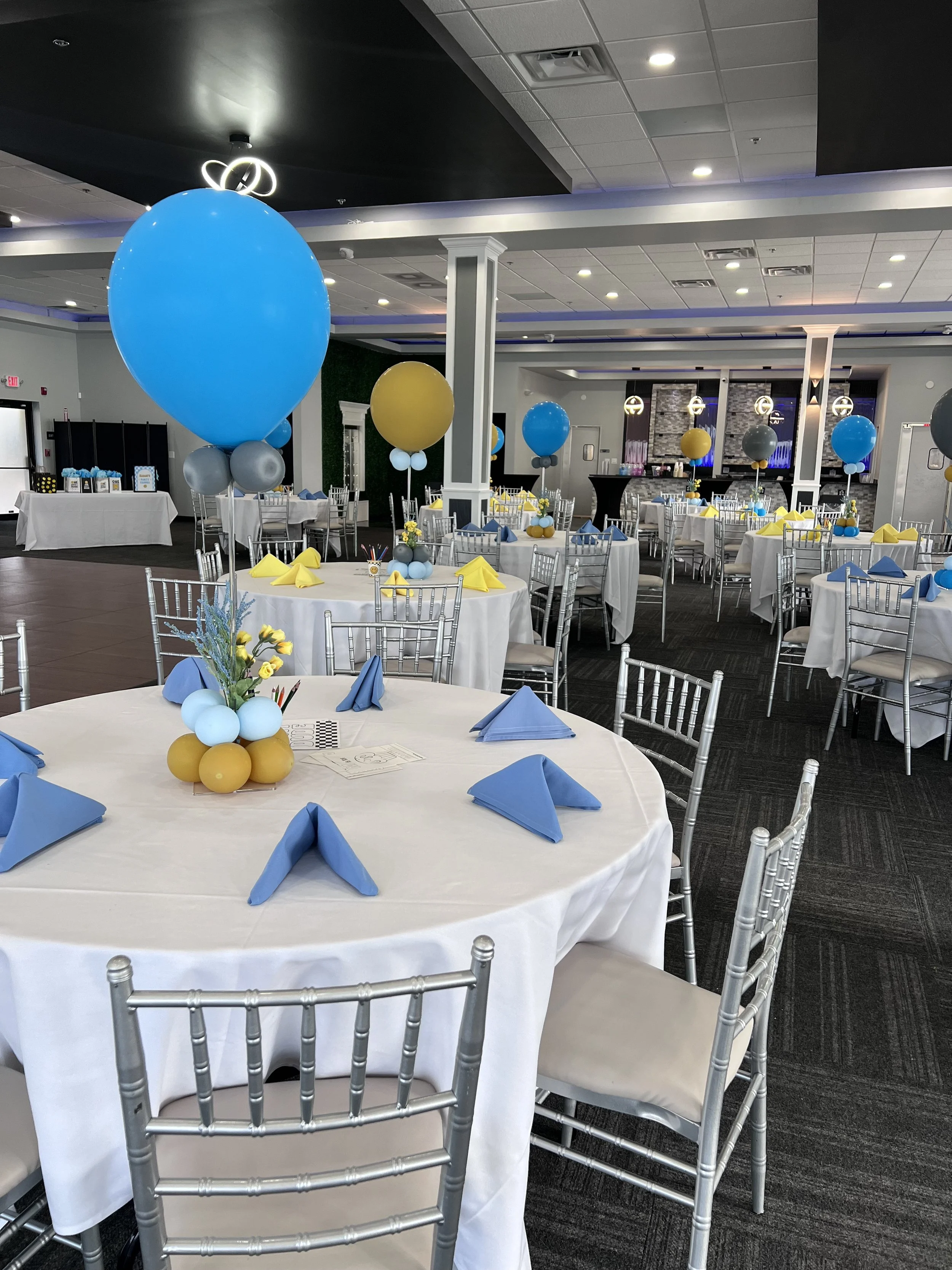 Party banquet hall decorated with blue, yellow, and gray balloons, round tables with white tablecloths, blue napkins, and floral centerpieces, silver chairs, and a bar area, set for a celebration or special event.