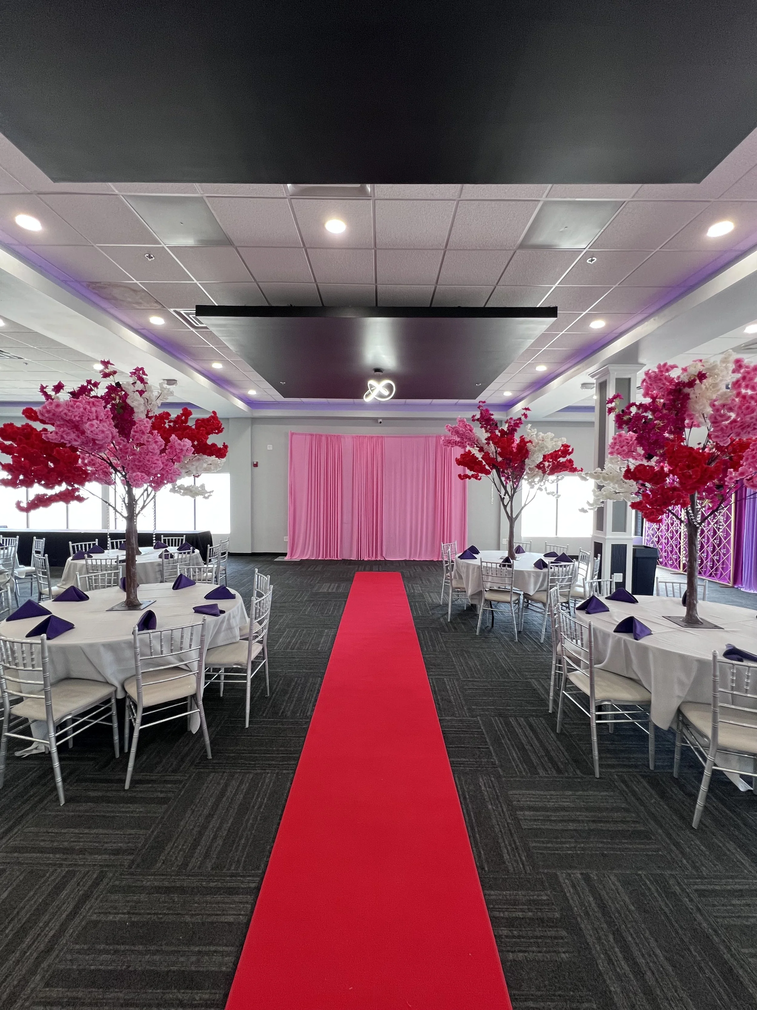Event space decorated with pink flowering trees, round tables with white tablecloths and navy napkins, and a pink curtain backdrop, with a red carpet leading to the stage area.