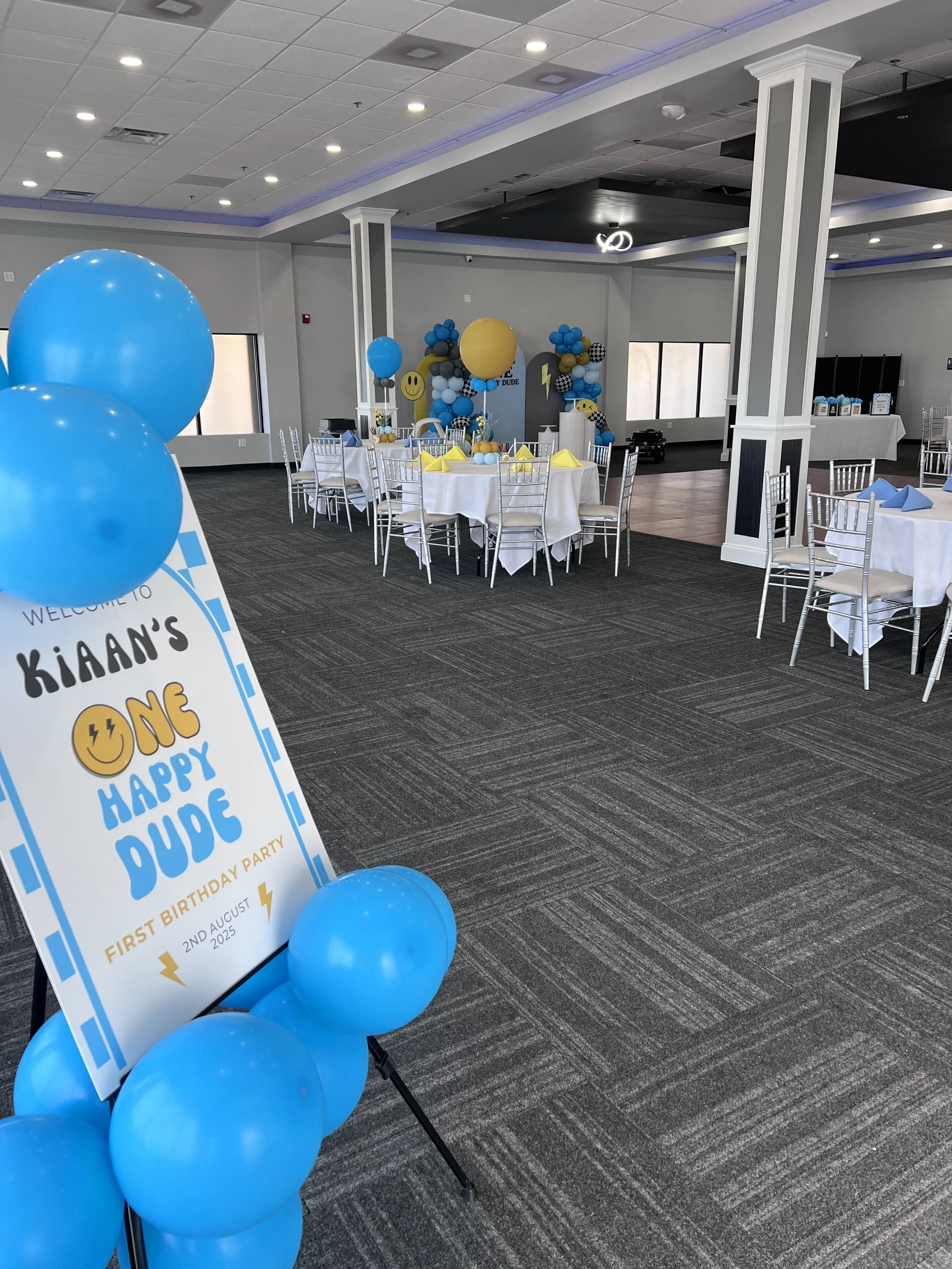 Decorated event space with tables, chairs, and blue and yellow balloons, celebrating a child's first birthday with themed banners and decorations.