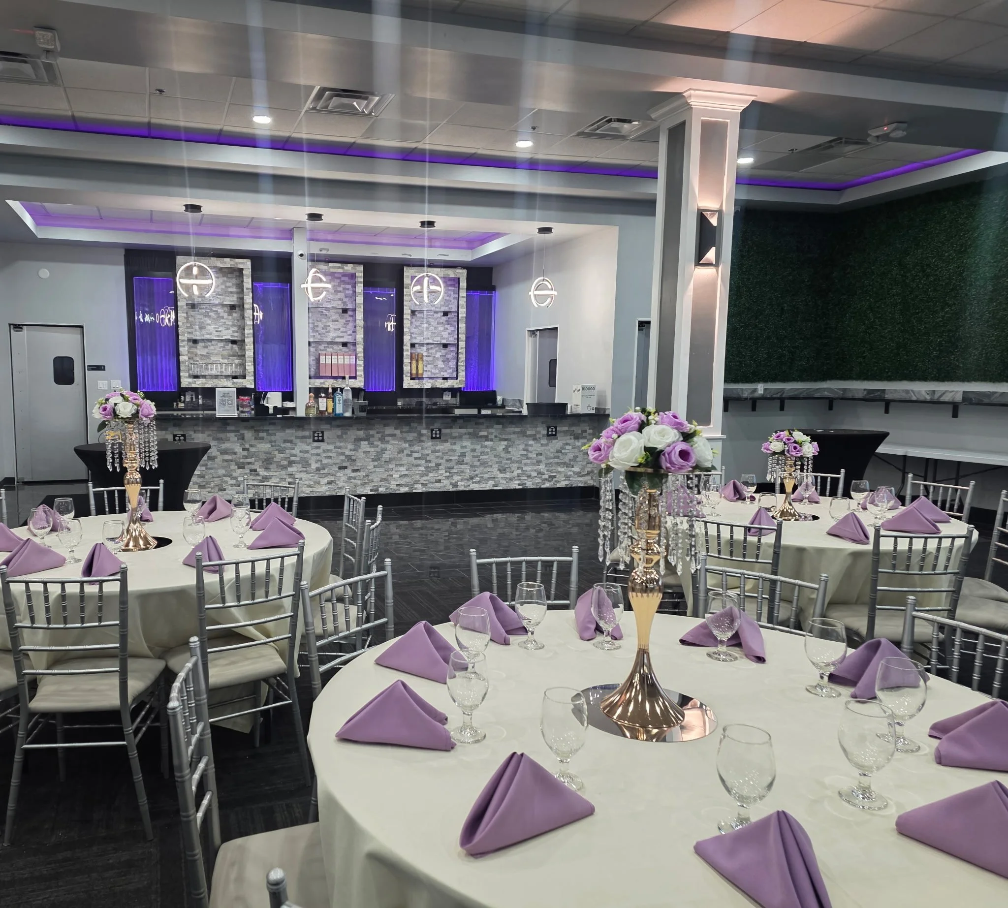 Interior of an event hall decorated with purple-themed table settings and elegant floral centerpieces, featuring a decorated bar area with purple lighting and modern pendant lights.