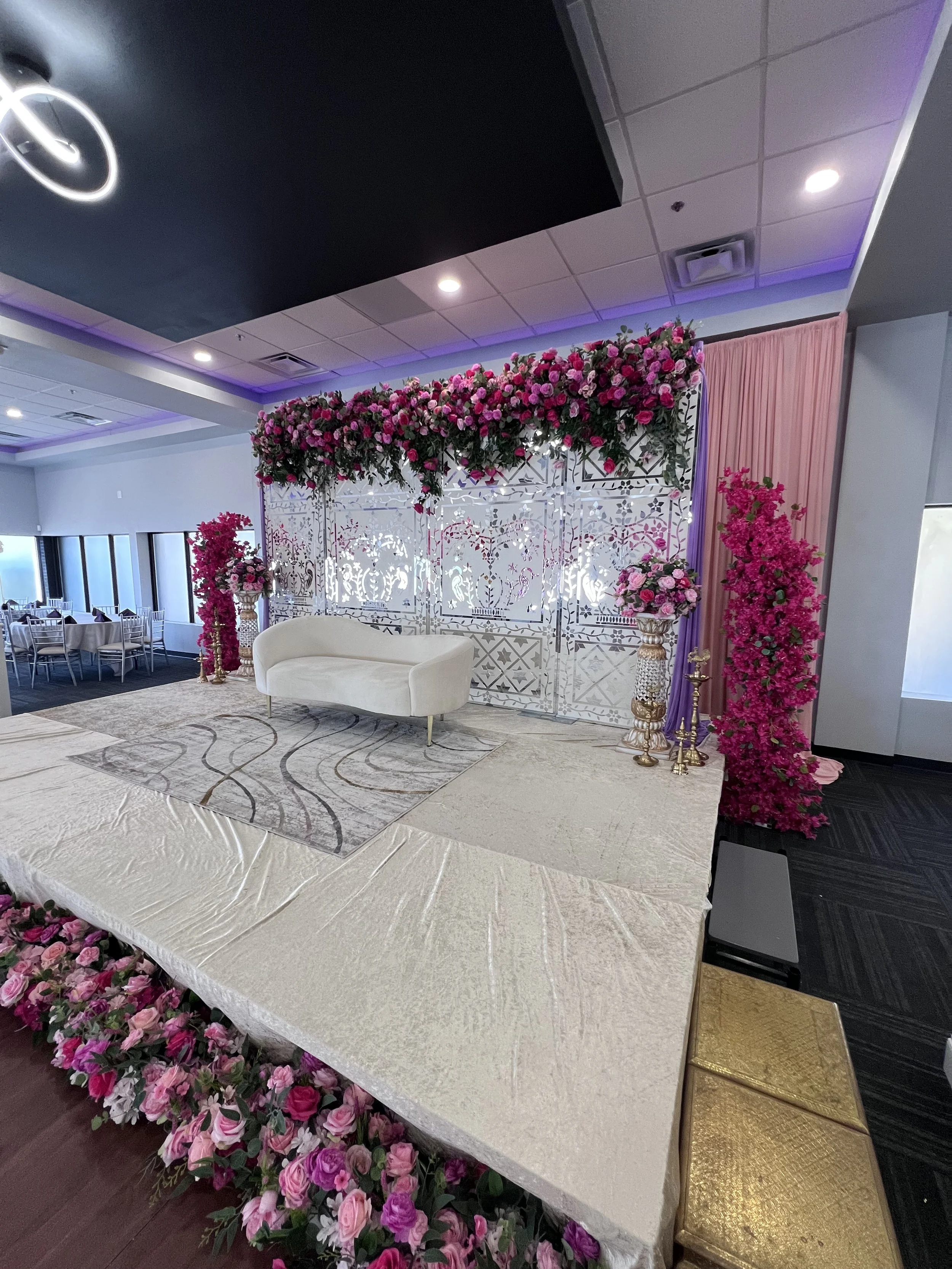 Decorative wedding stage with a floral arch of pink roses, white lattice backdrop, white furniture, and pink and gold accents.