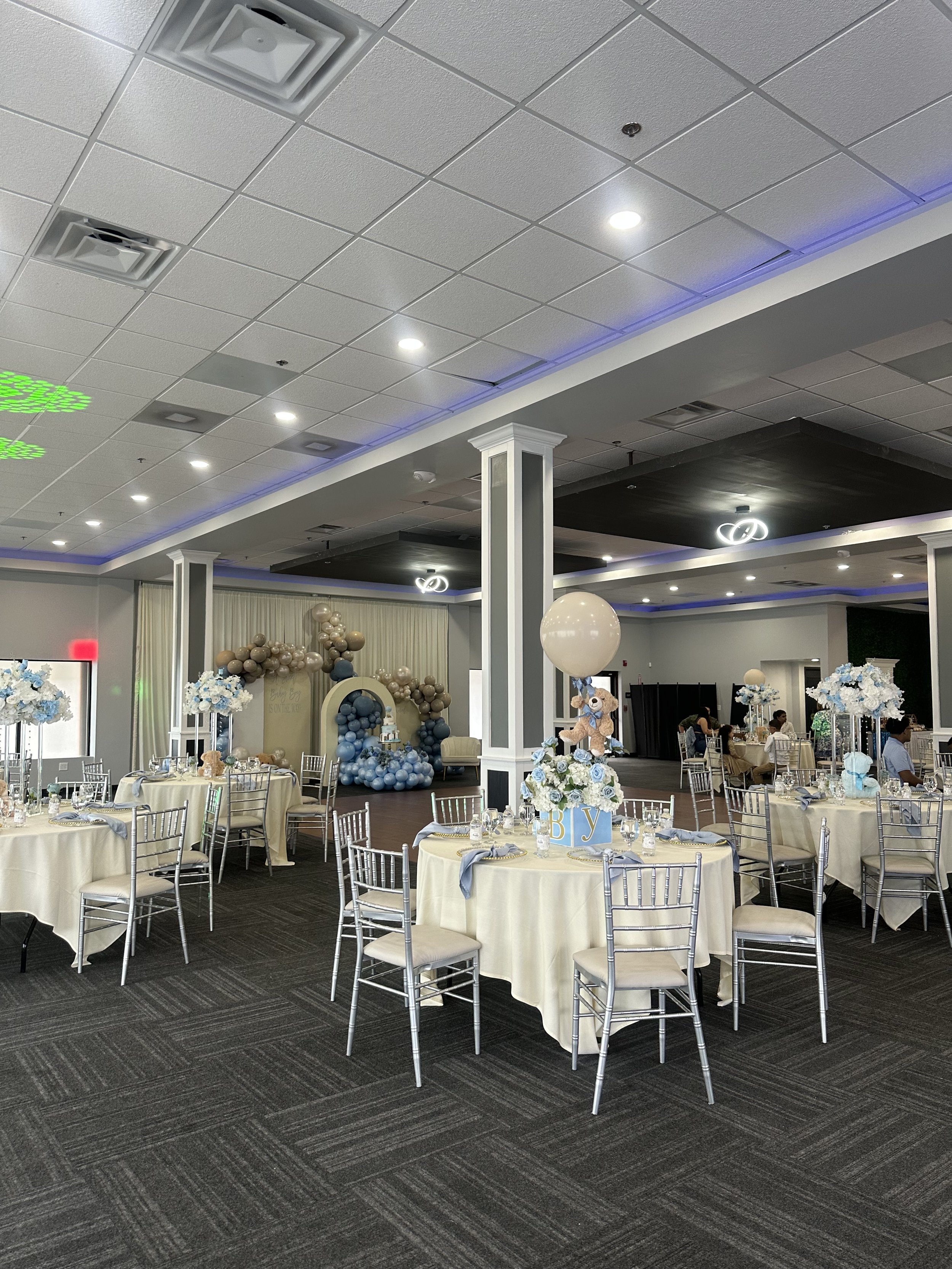 Decorated banquet hall with tables set for a celebration, featuring balloon arrangements, floral centerpieces, and a sign that says 'Baby'.