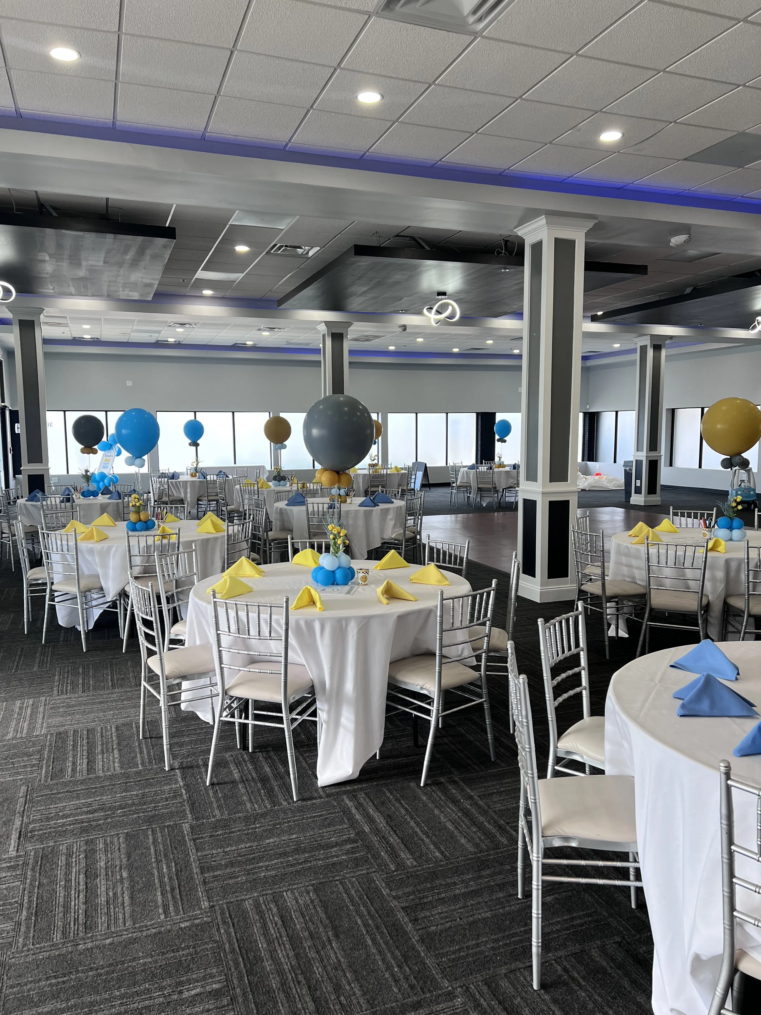 A banquet hall decorated for a celebration, featuring round tables with white tablecloths, yellow and blue napkins, and balloon centerpieces in blue, yellow, black, and gray. The hall has large windows, a patterned carpet, and modern ceiling lighting.