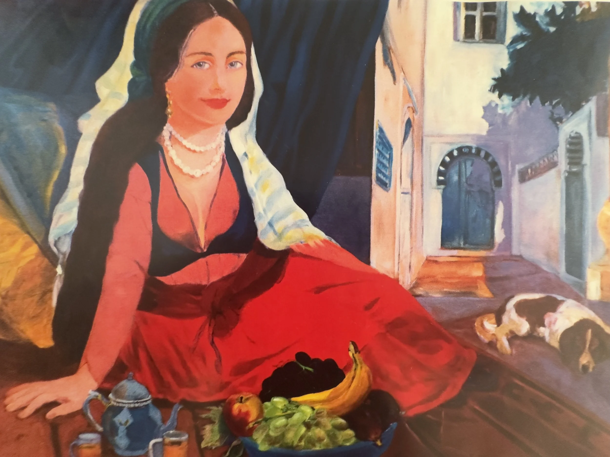 A woman with black hair and pale skin wearing red lipstick, a pearl necklace, and earrings, sitting in front of a dark curtain, with colorful artwork of buildings and a large green plant in the background. She is dressed in a red dress with black acc