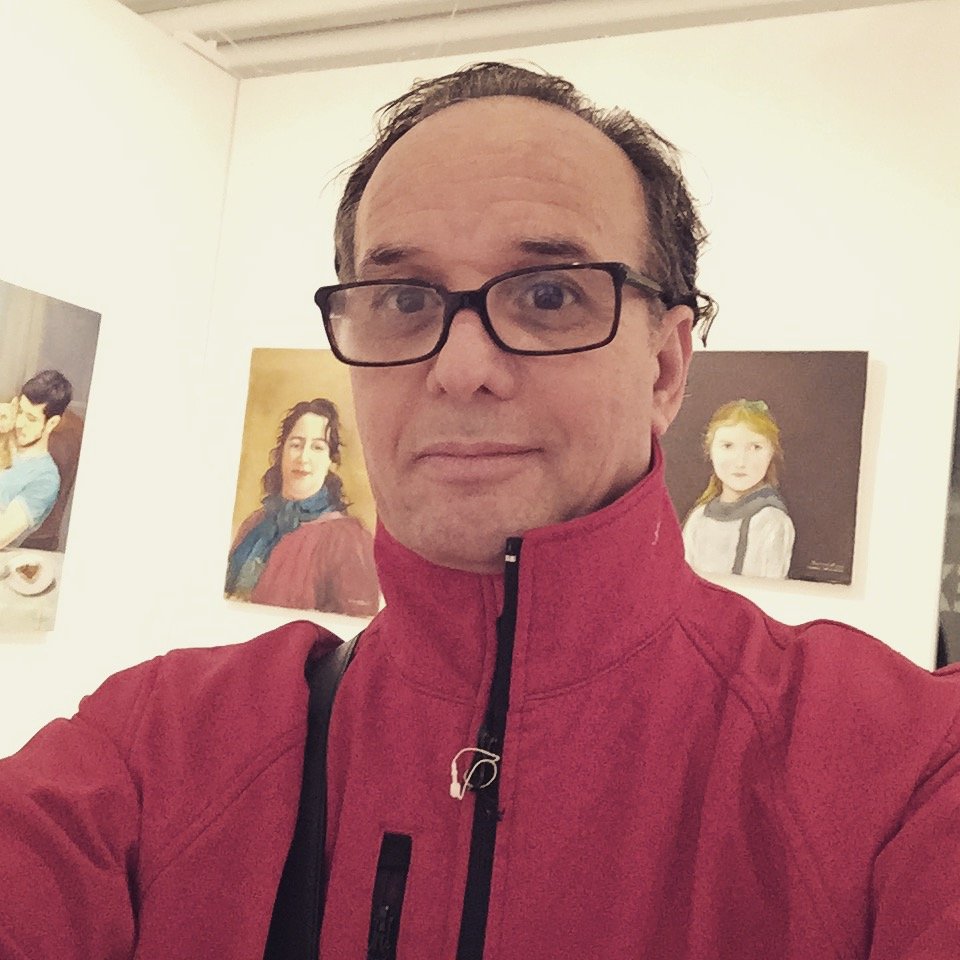 A man wearing glasses and a red jacket takes a selfie in an art gallery. Behind him, there are two portrait paintings of women on the wall.