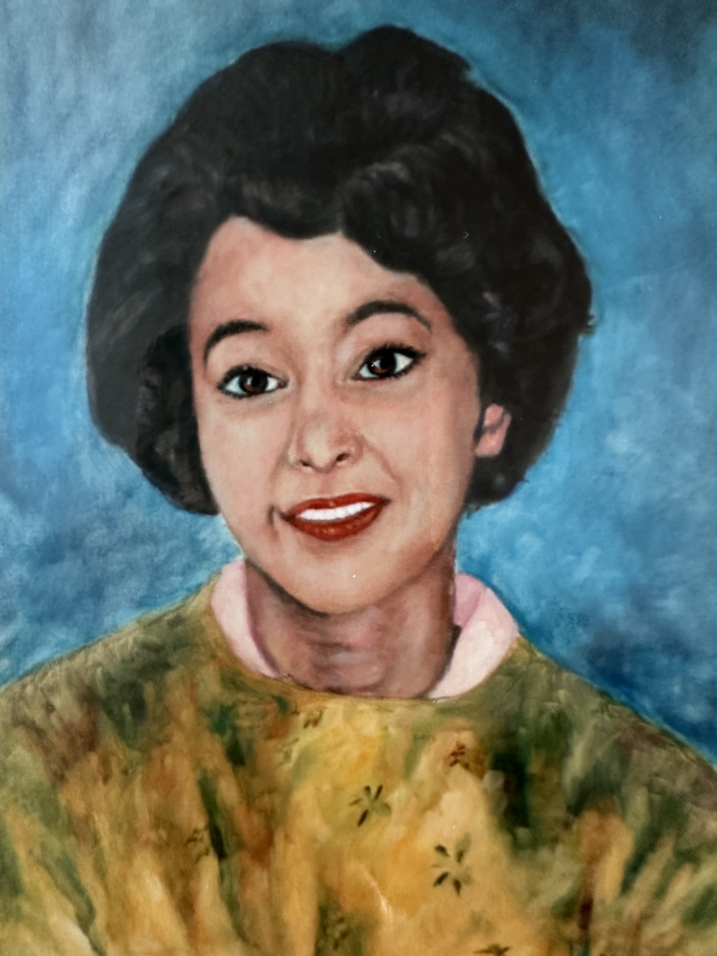A woman with short dark hair, wearing a colorful patterned blouse, smiling against a blue background.