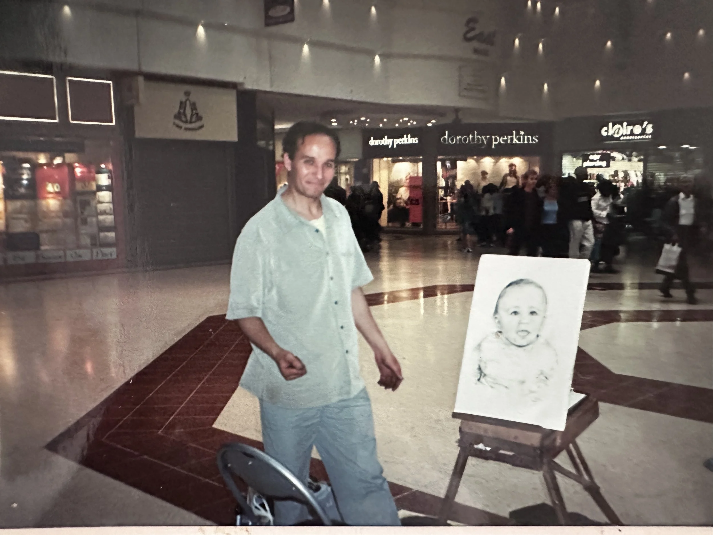 A young man in a light green shirt standing in a shopping mall, with a portrait of a baby on an easel and some store signs, including Dorothy Perkins and Claire's, visible in the background.