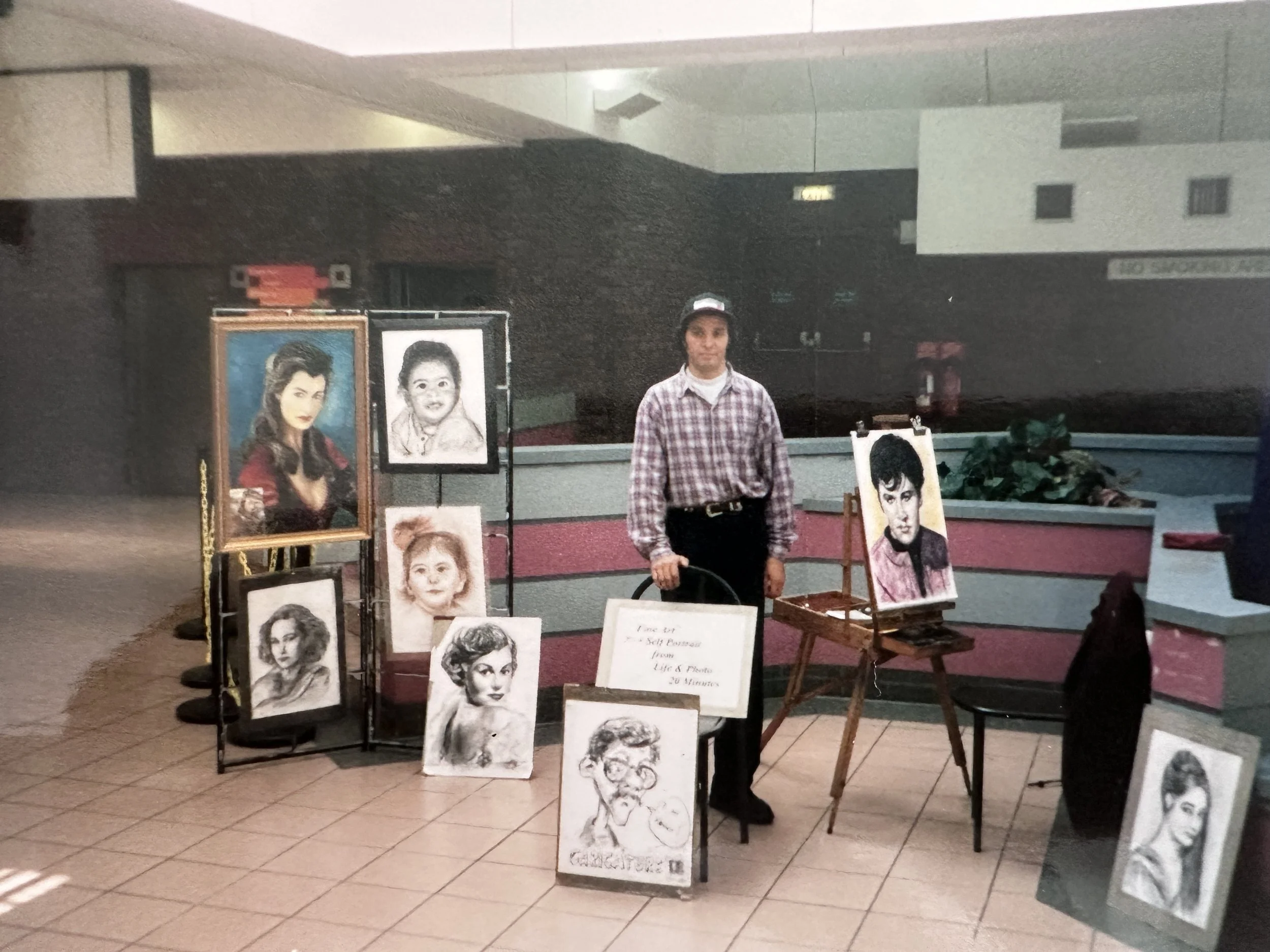 An indoor art display features portraits of women and children, including a realistic colored painting of a woman with dark hair and a black-and-white sketch of a smiling child. Several artworks are arranged on easels and stands, with a person standi