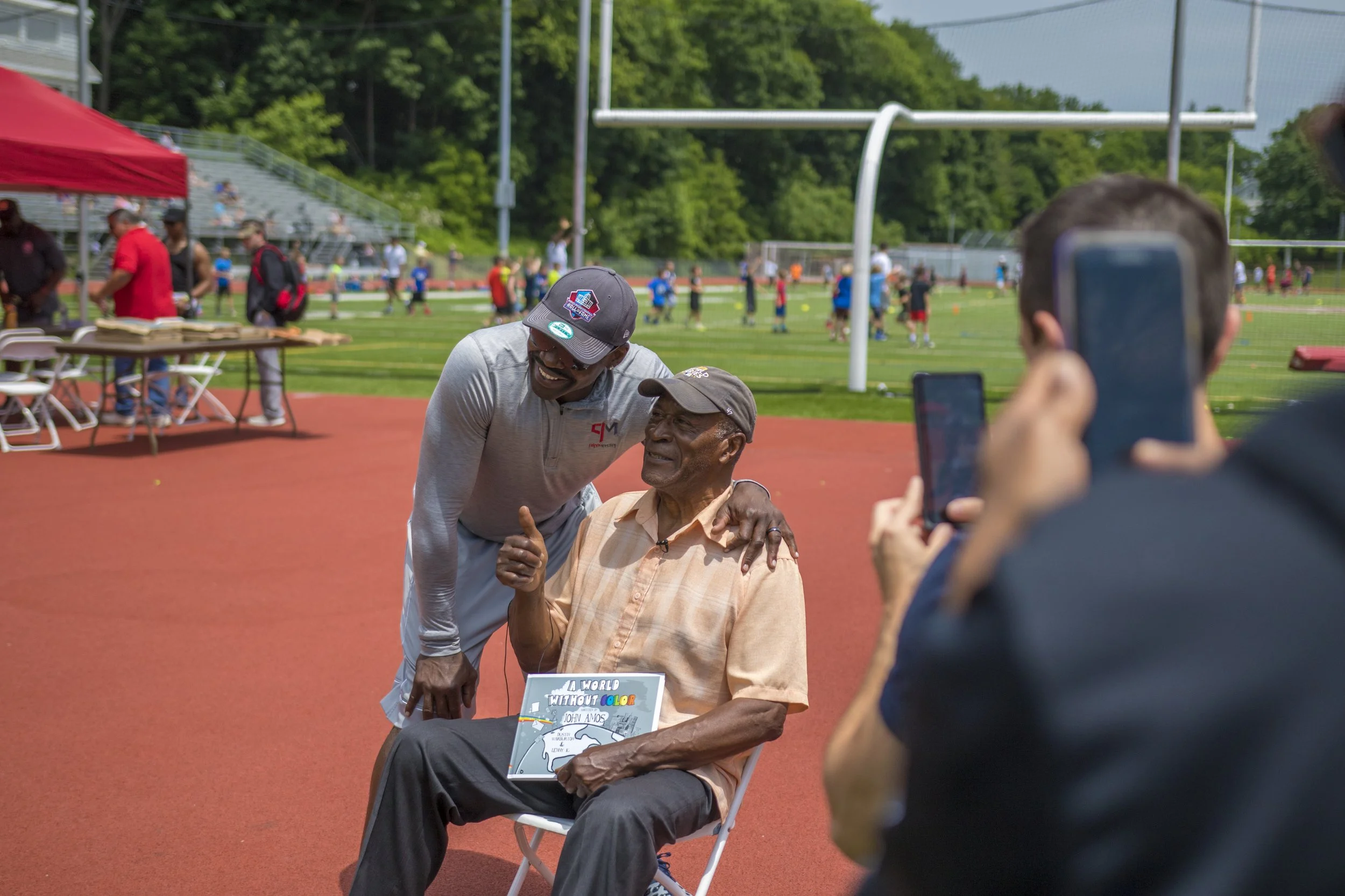 Actor John Amos holding his book A World Without Color while speaking with Dallas Cowboys legend Michael Irvin at an outdoor event.