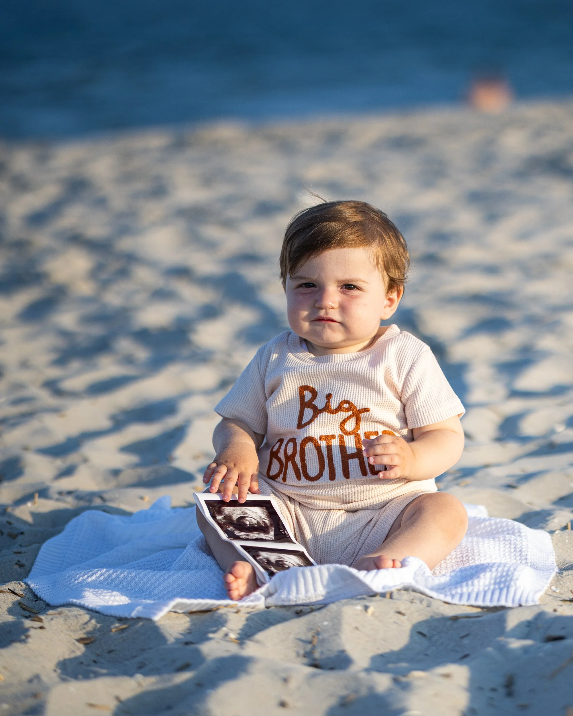 A young child sitting on a white blanket on a sandy beach, holding an ultrasound image, wearing a beige shirt with the words 'Big Brother' in brown, with the ocean and a blurry boat in the background.
